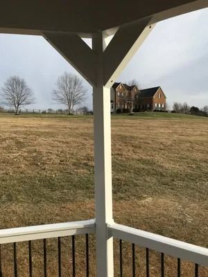 View from a white porch frame overlooking a brown field towards a brick house on a hill under a cloudy sky.