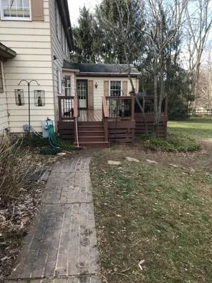 A house with a brick pathway leading to a deck with stairs, surrounded by grass and trees.