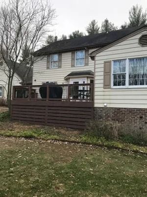 Brown deck attached to a beige two-story house, with a grill and trees in the yard.