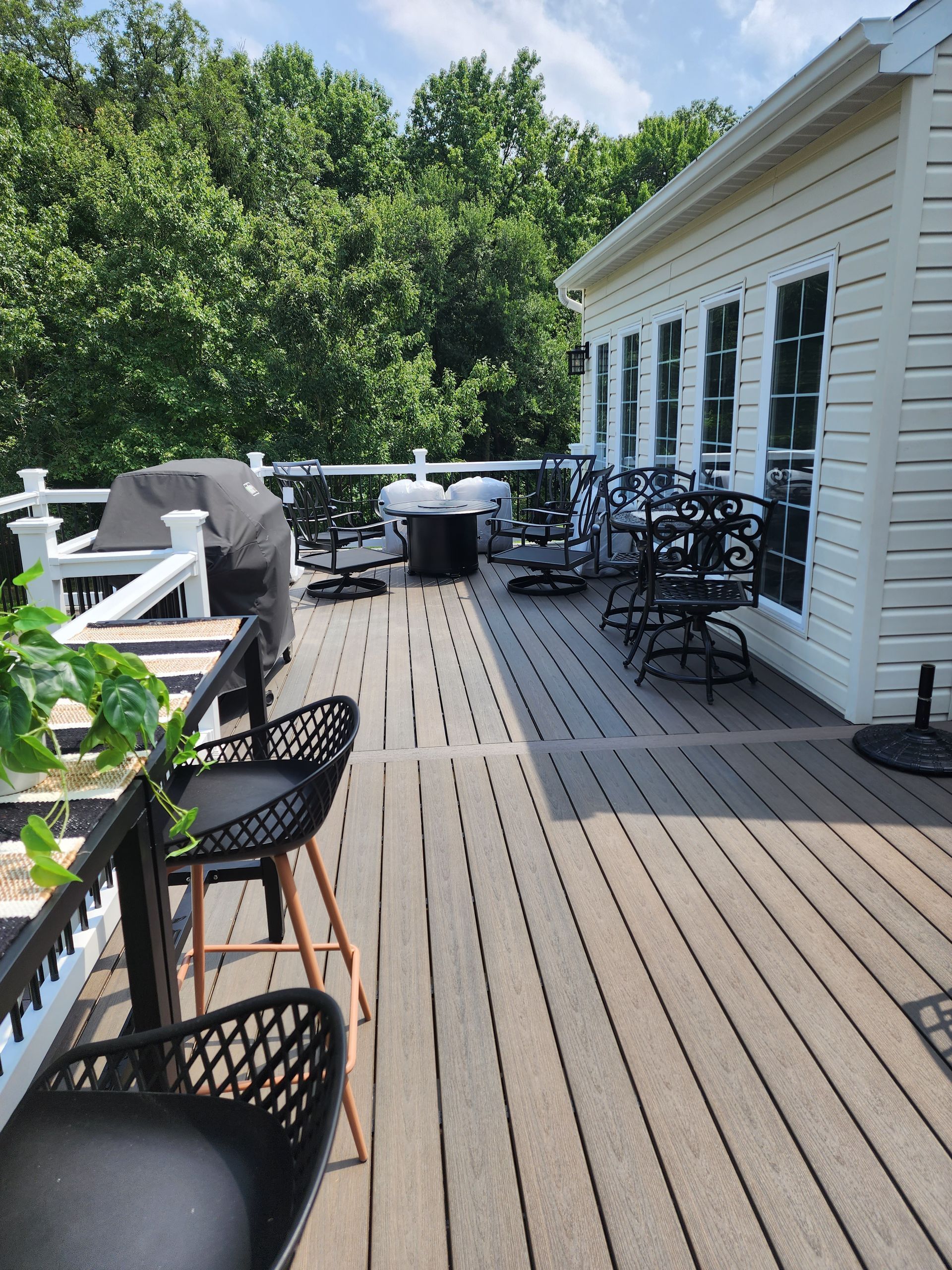 Deck with black chairs, grill, and round table; next to a white house, backed by trees.