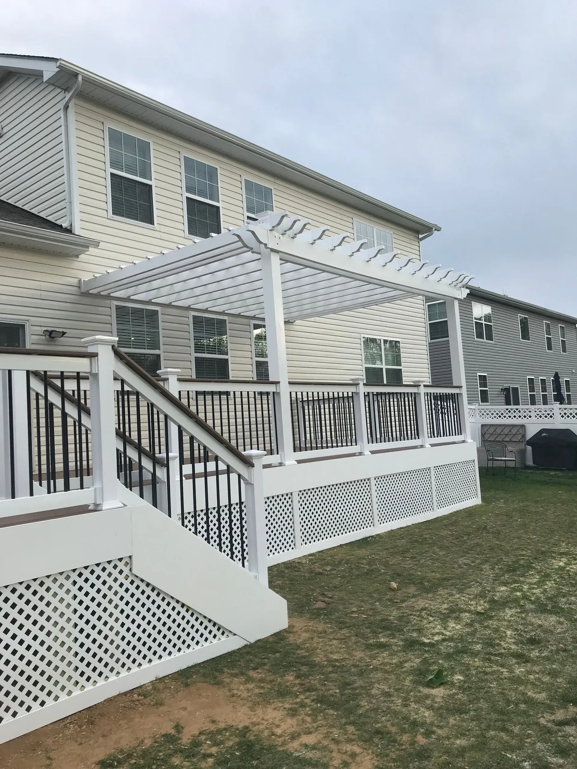 White deck with pergola attached to a two-story beige house; black railing and lattice detailing.
