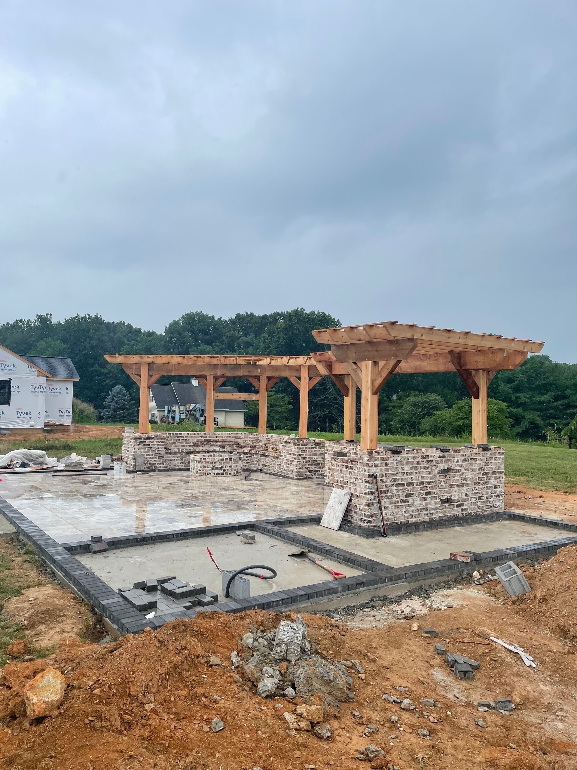 Construction site with pergola frame over brick structures, concrete patio, cloudy sky.