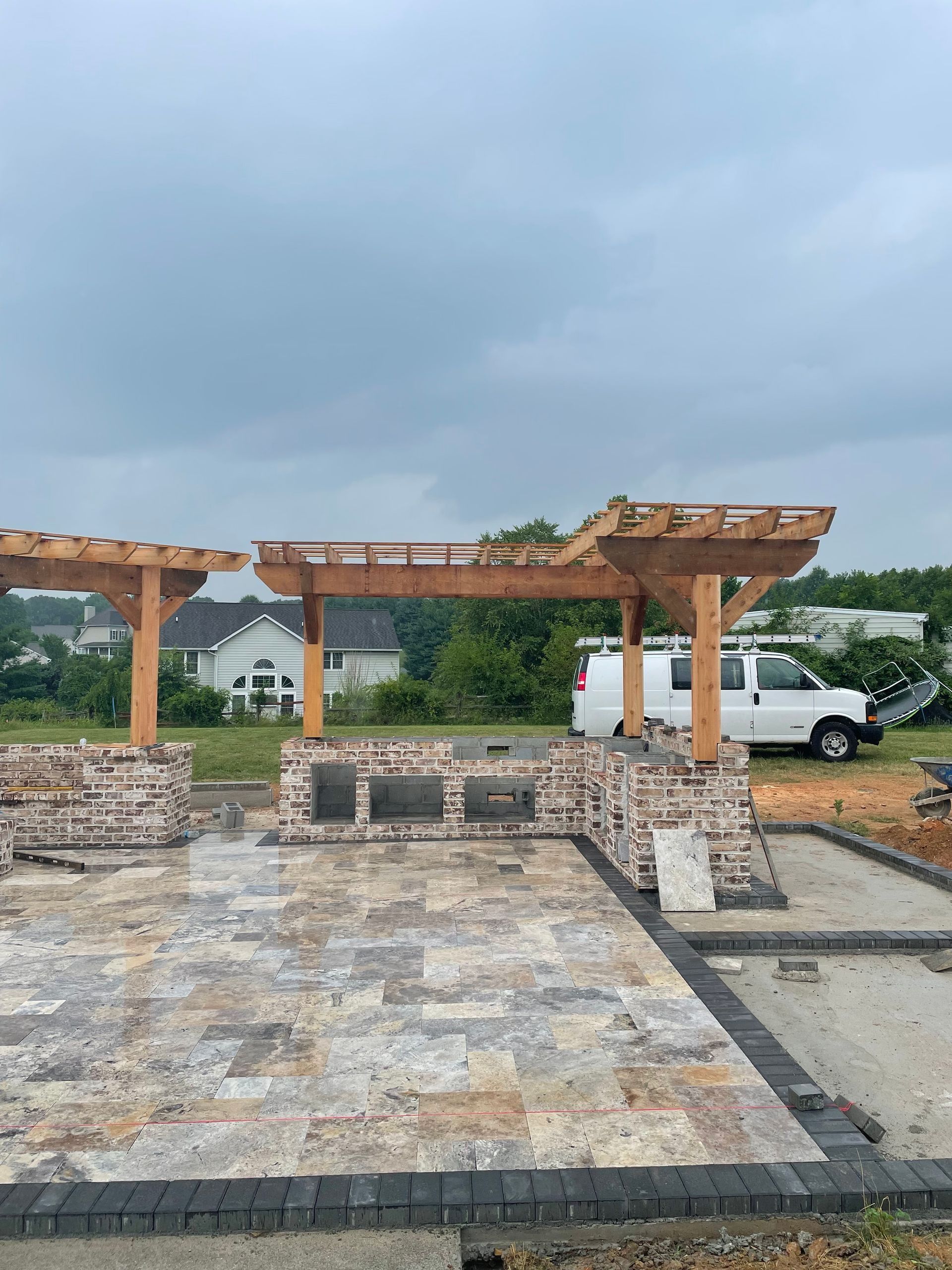 Outdoor kitchen under construction with pergola frame, stone, and patio. White van parked nearby.