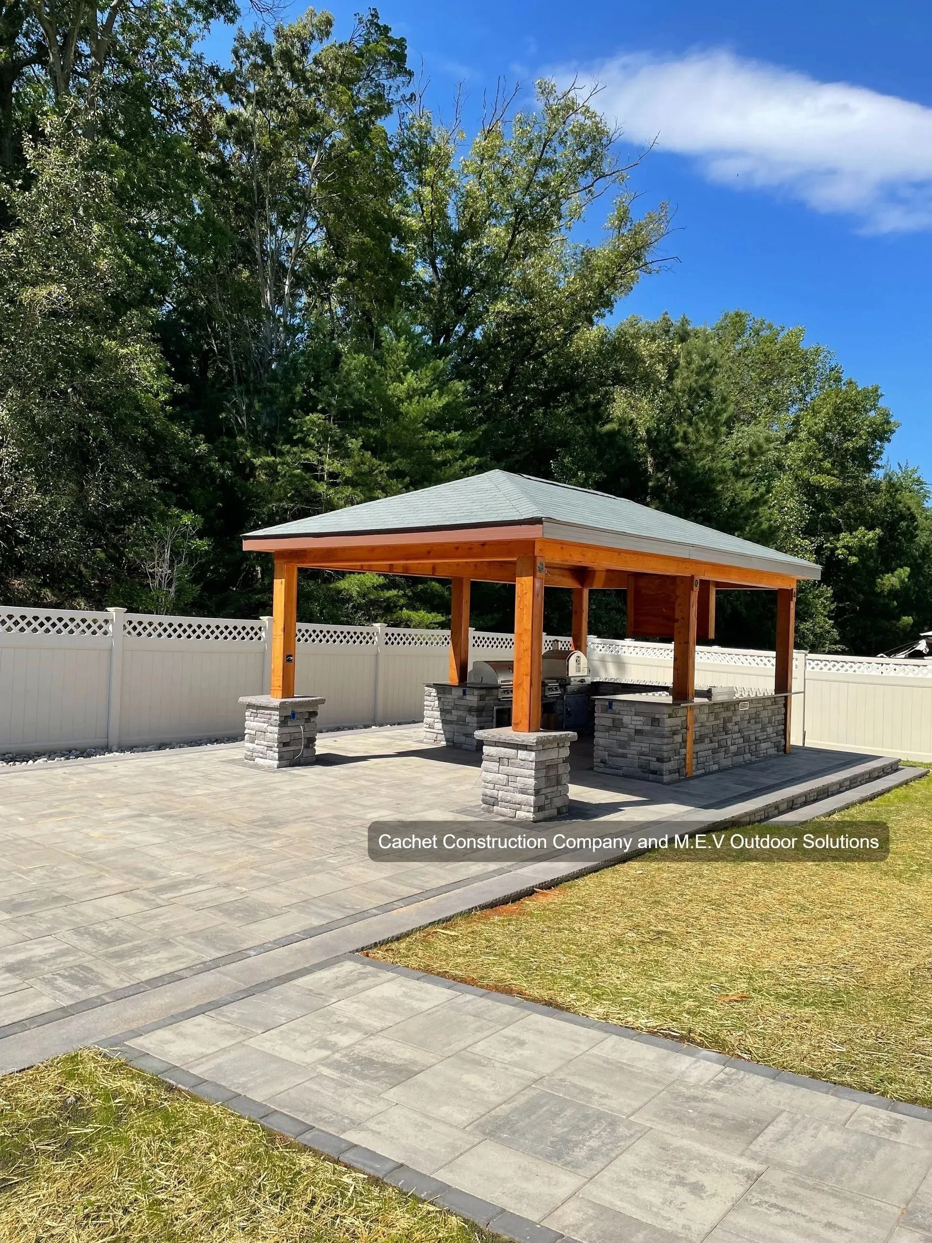 Outdoor gazebo with a stone base and wooden frame, on a paved patio. Green grass and trees in the background.