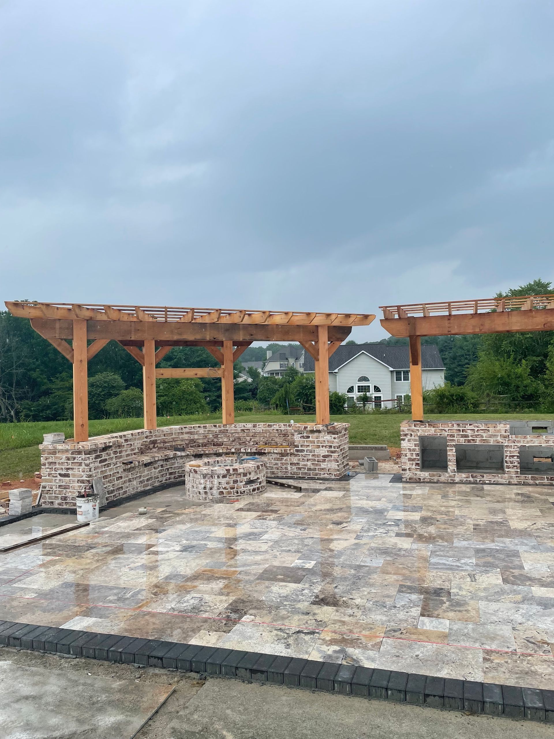 Outdoor kitchen under construction with wood pergola, brick base, and paver patio. Overcast sky.
