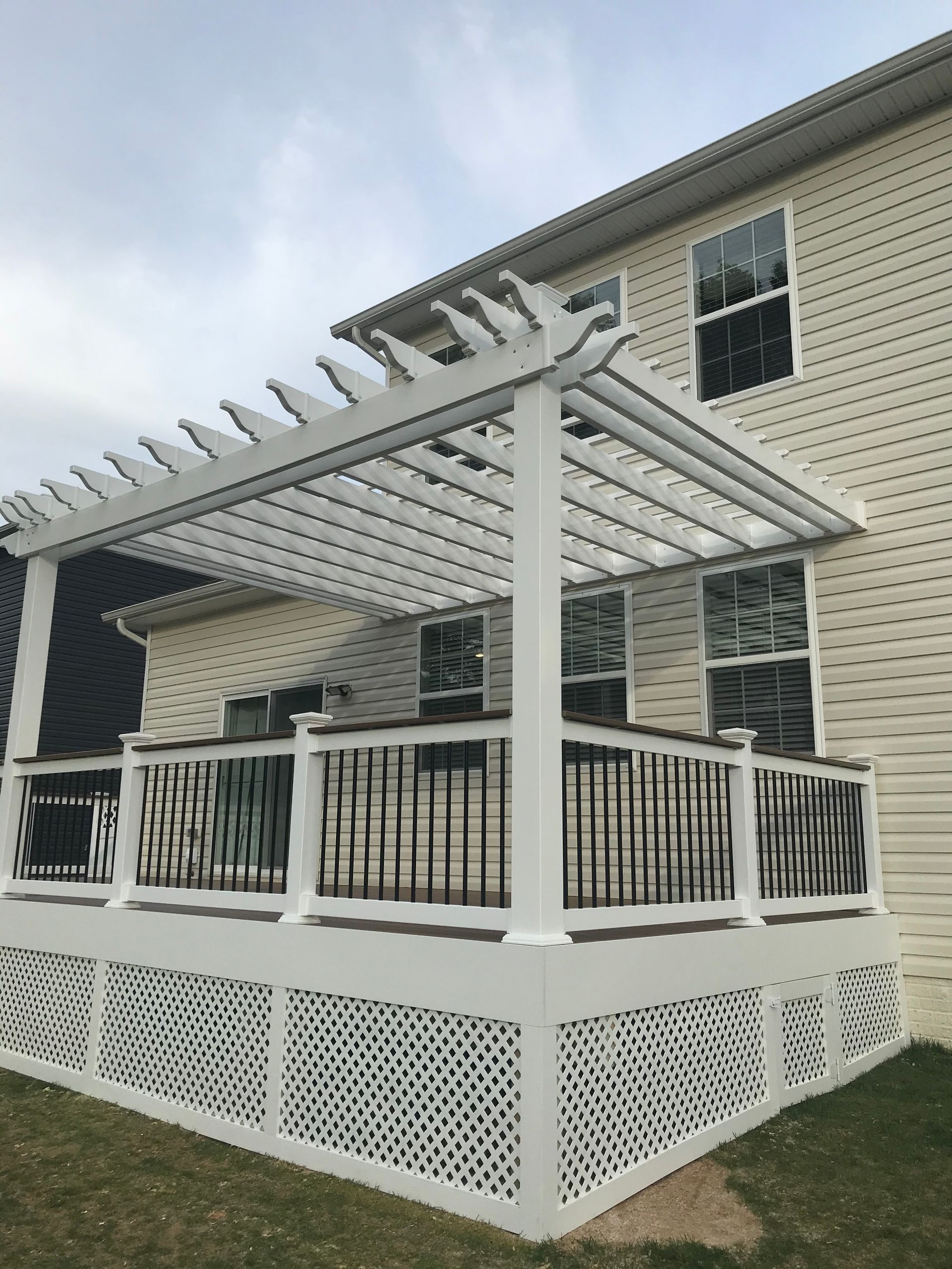 White pergola over a deck with lattice siding attached to a tan house, against a cloudy sky.