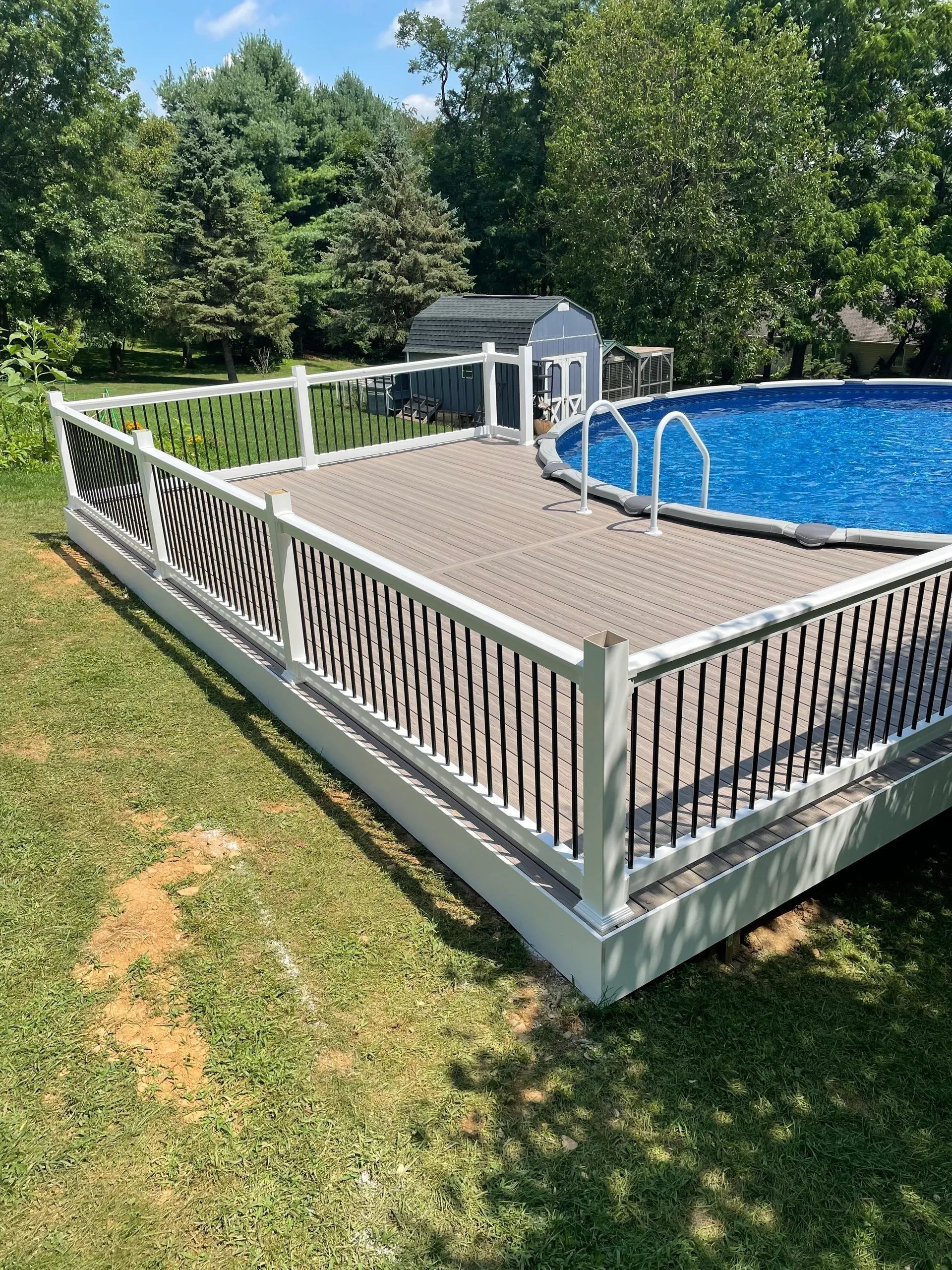 Above-ground pool with a deck, white and black railing, blue water, and surrounding green grass and trees.