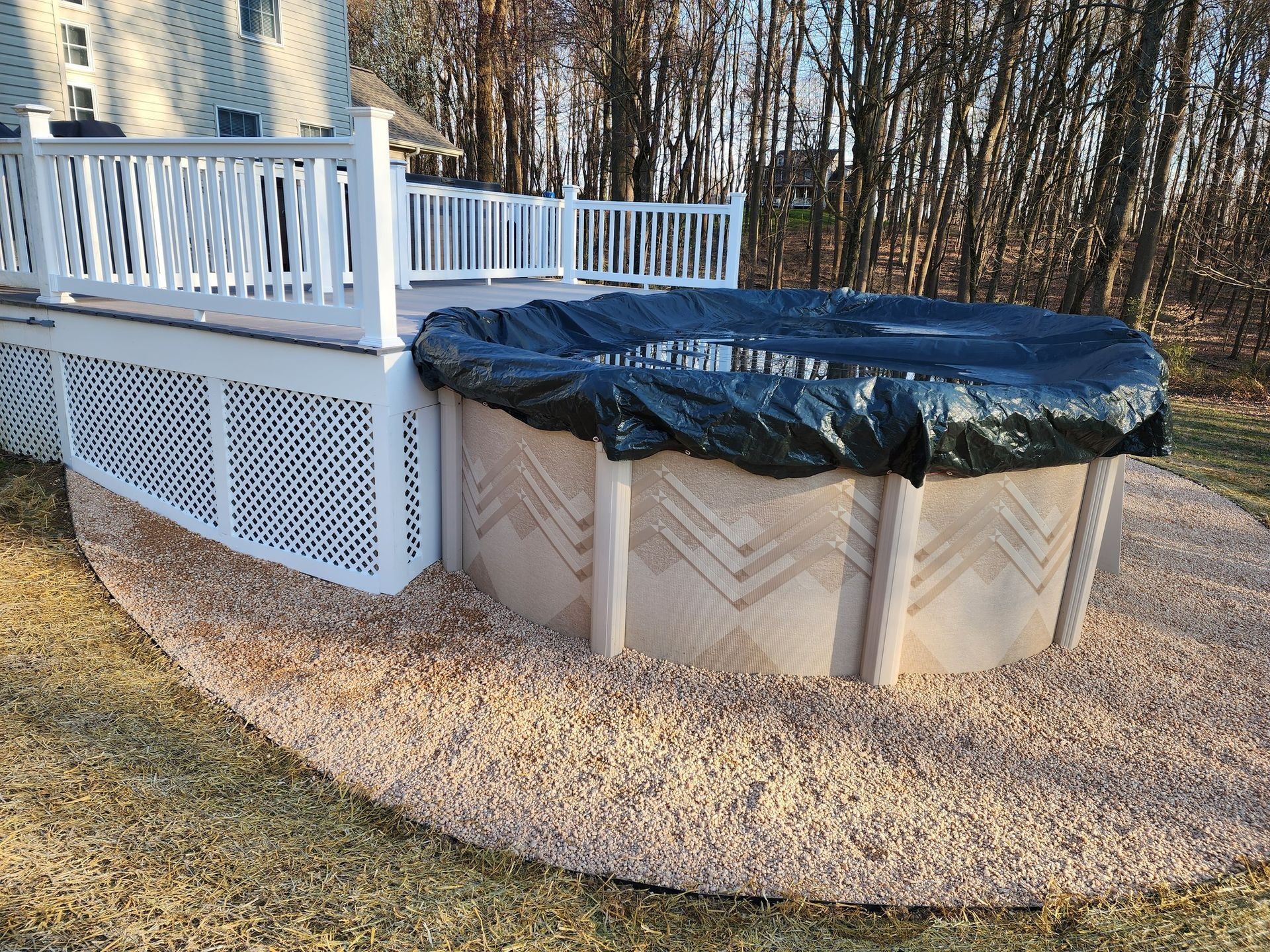 Above-ground pool covered with black tarp, next to a white deck. Surrounded by tan gravel, on grassy lawn.