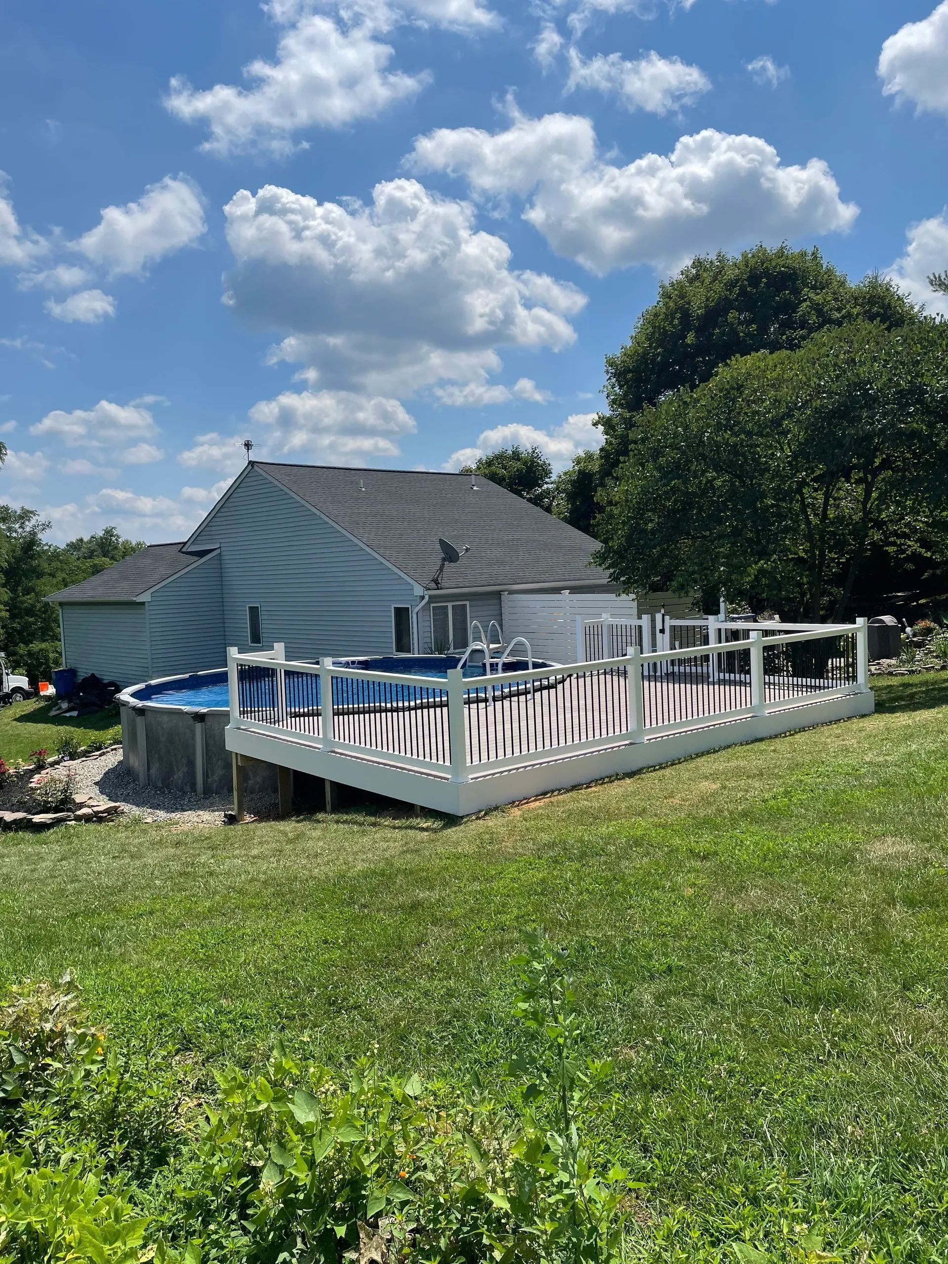 Backyard with above-ground pool and white deck, set against a house and blue sky with clouds. Green grass in the foreground.