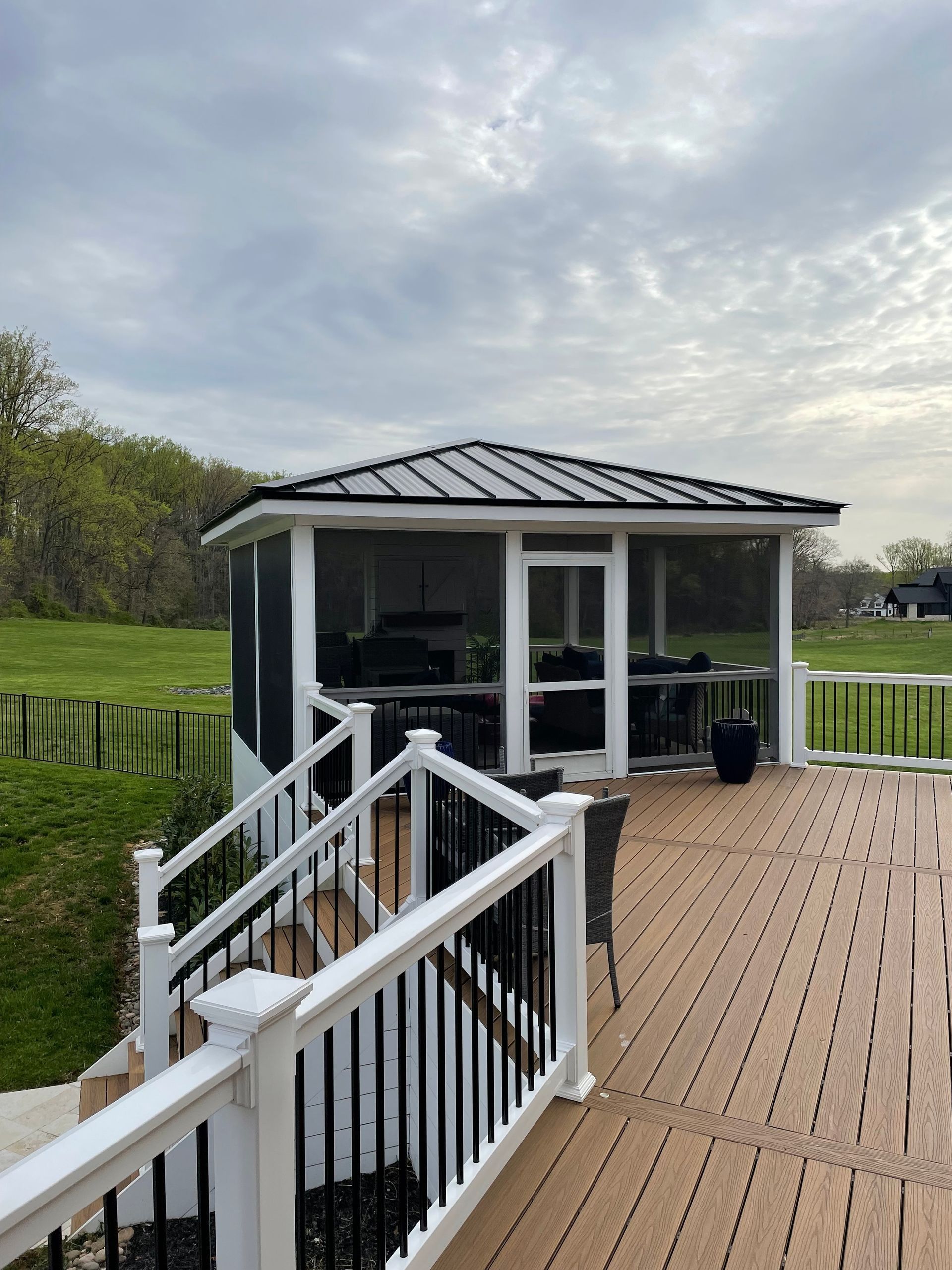 Screened gazebo on a wooden deck, black railing, grassy hill in background, cloudy sky.