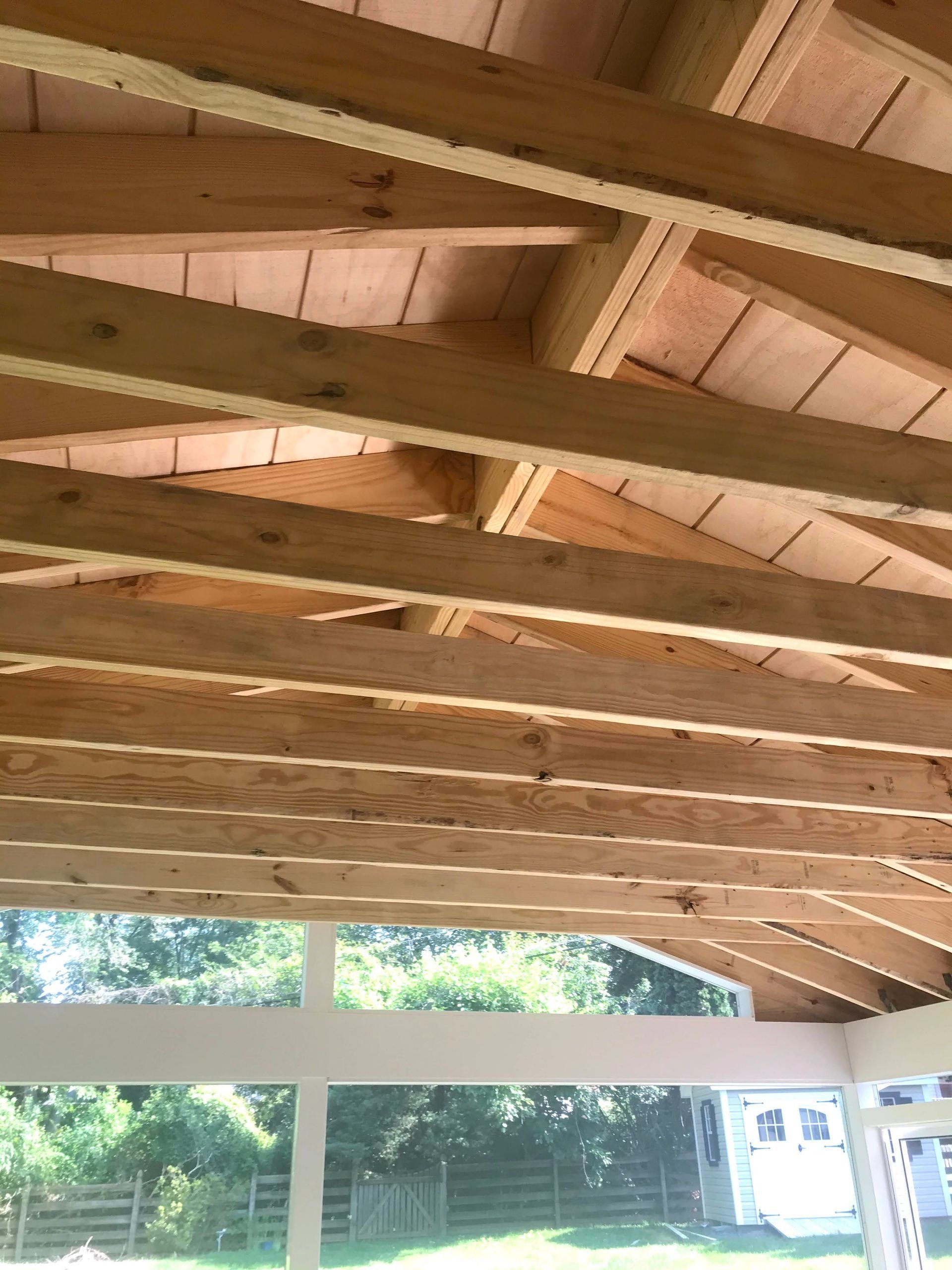 Wooden ceiling frame with exposed rafters and supporting beams over a porch overlooking a backyard.