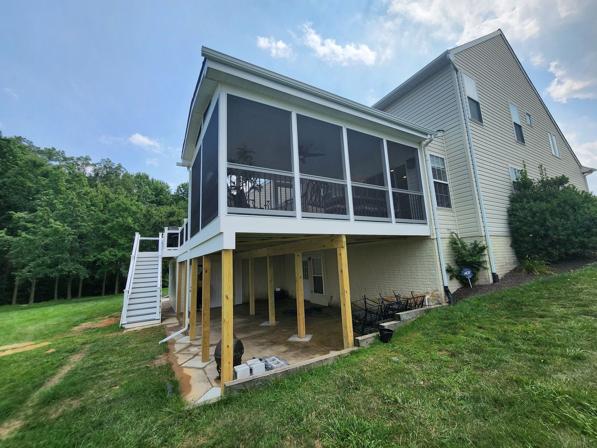 Screened porch with white trim and deck, built on a grassy hillside, attached to a two-story beige house.