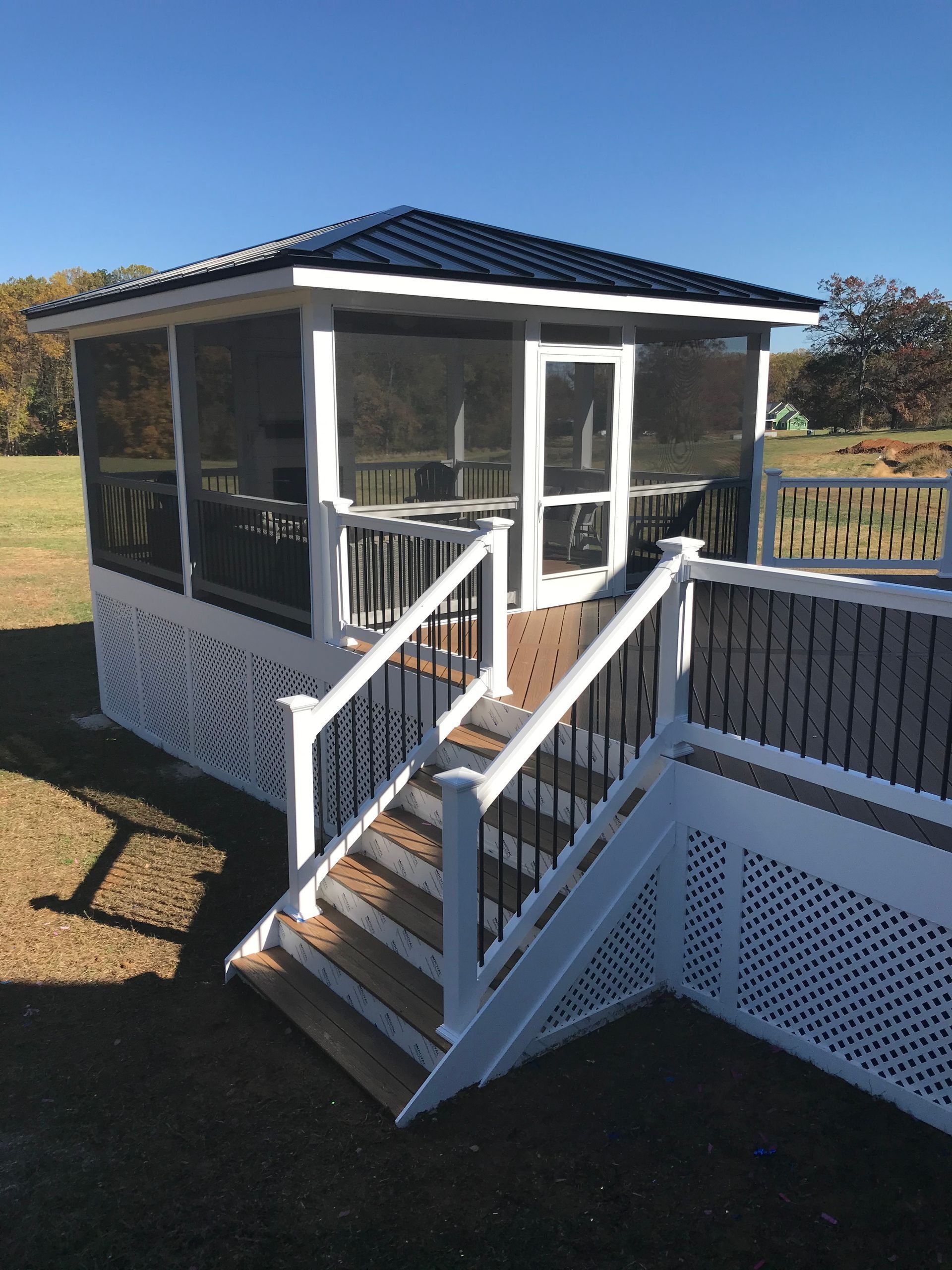Screened-in gazebo with stairs. White frame, black metal roof, black railings, and brown deck. Outdoors, sunny day.