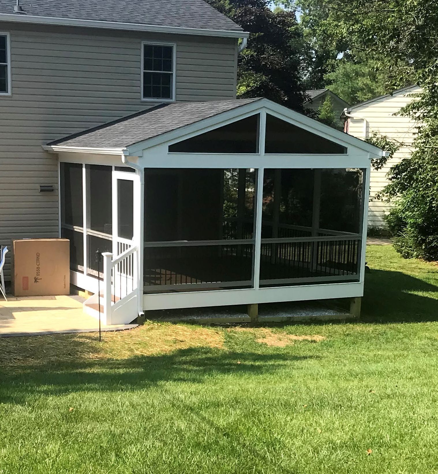 Screened-in porch attached to a light-colored house with a dark roof. Green grass and trees surround the structure.