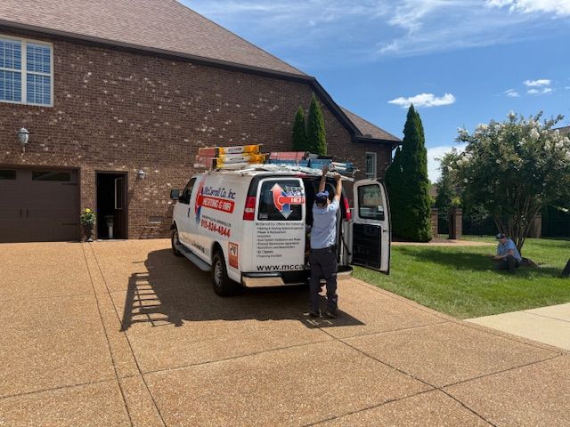 A man with a service van parked in a driveway. He's reaching for something on the roof. Brick house on a sunny day.