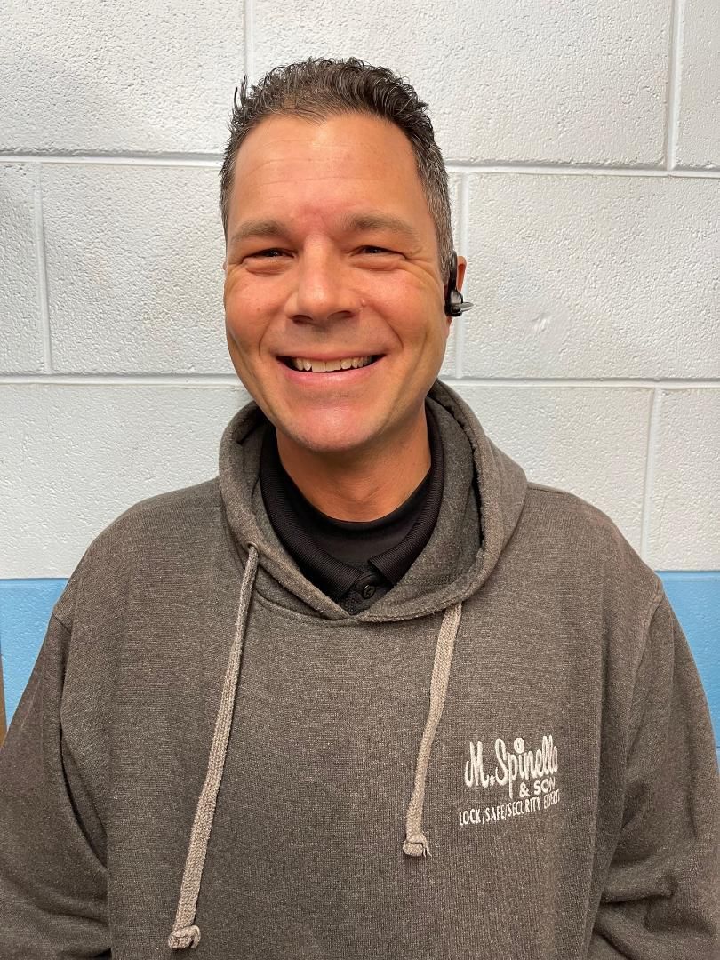 Man with graying hair in a gray hoodie smiles at the camera, wearing an earpiece; background is a light blue and white wall.
