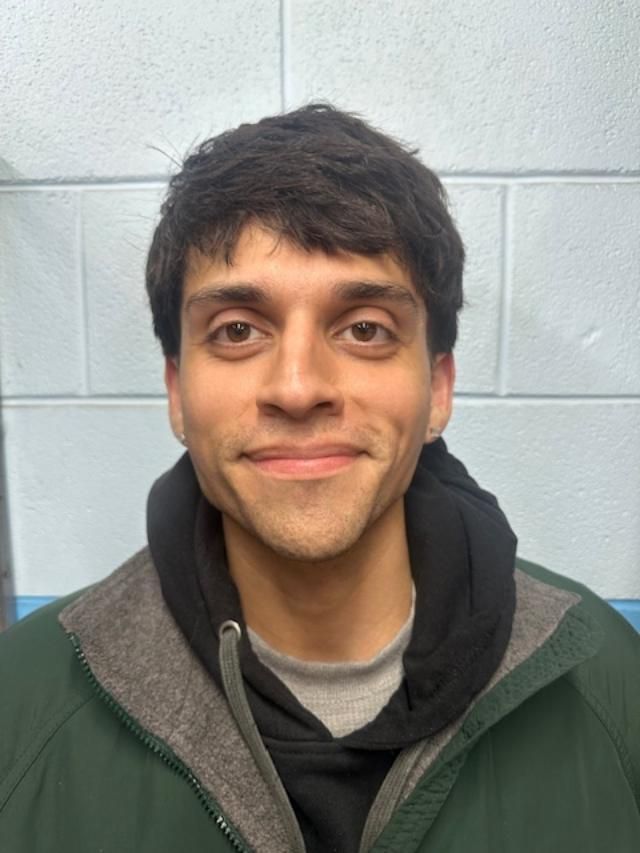 Man with dark hair and brown eyes, wearing a green jacket over a grey hoodie, smiling slightly in front of a blue tiled wall.