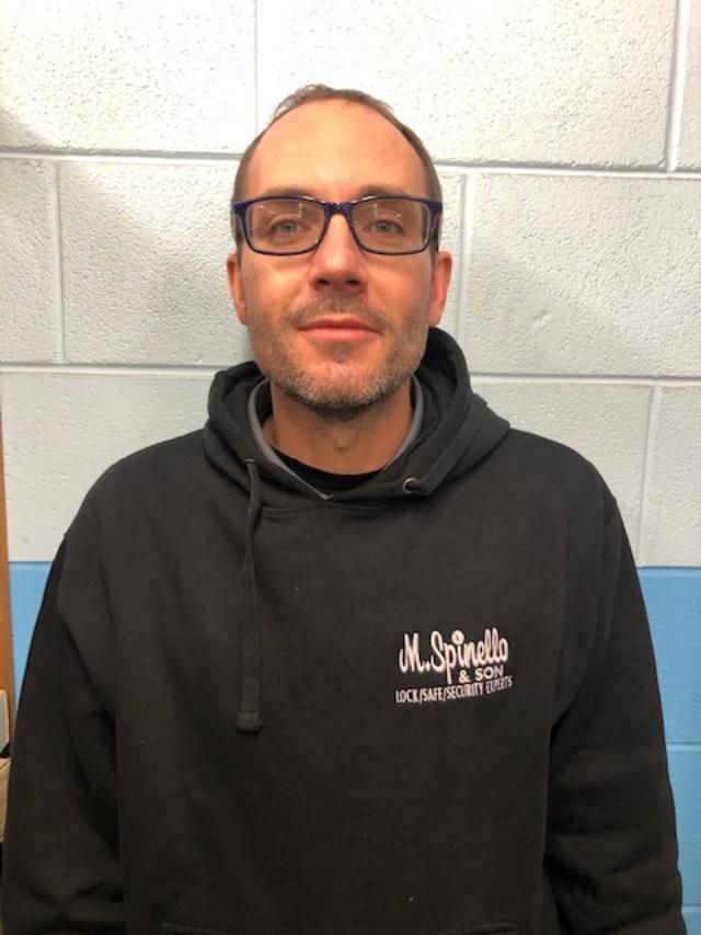 Man wearing glasses and a black hoodie with logo, standing in front of a blue and white wall.