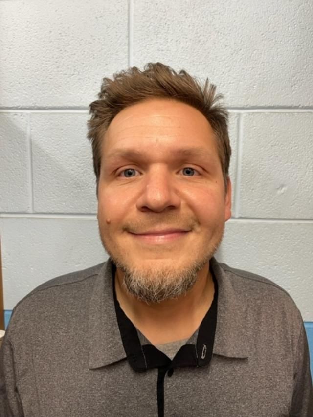 Man with short brown hair and beard smiles, wearing a gray collared shirt, in front of a white and blue brick wall.
