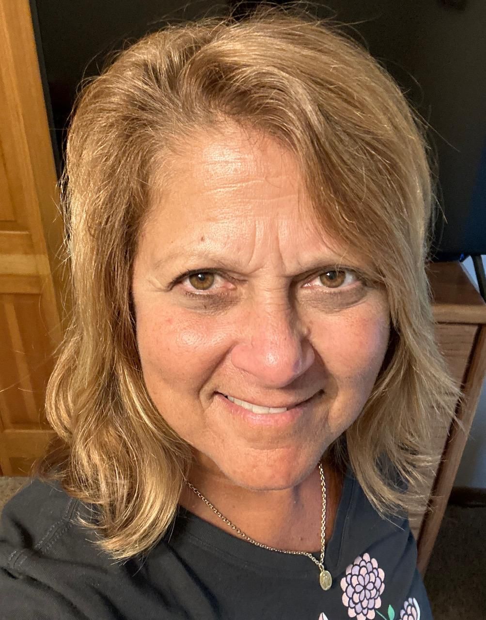 Woman with shoulder-length blonde hair, smiling at camera. Wearing a dark shirt with flower design. Indoor setting.