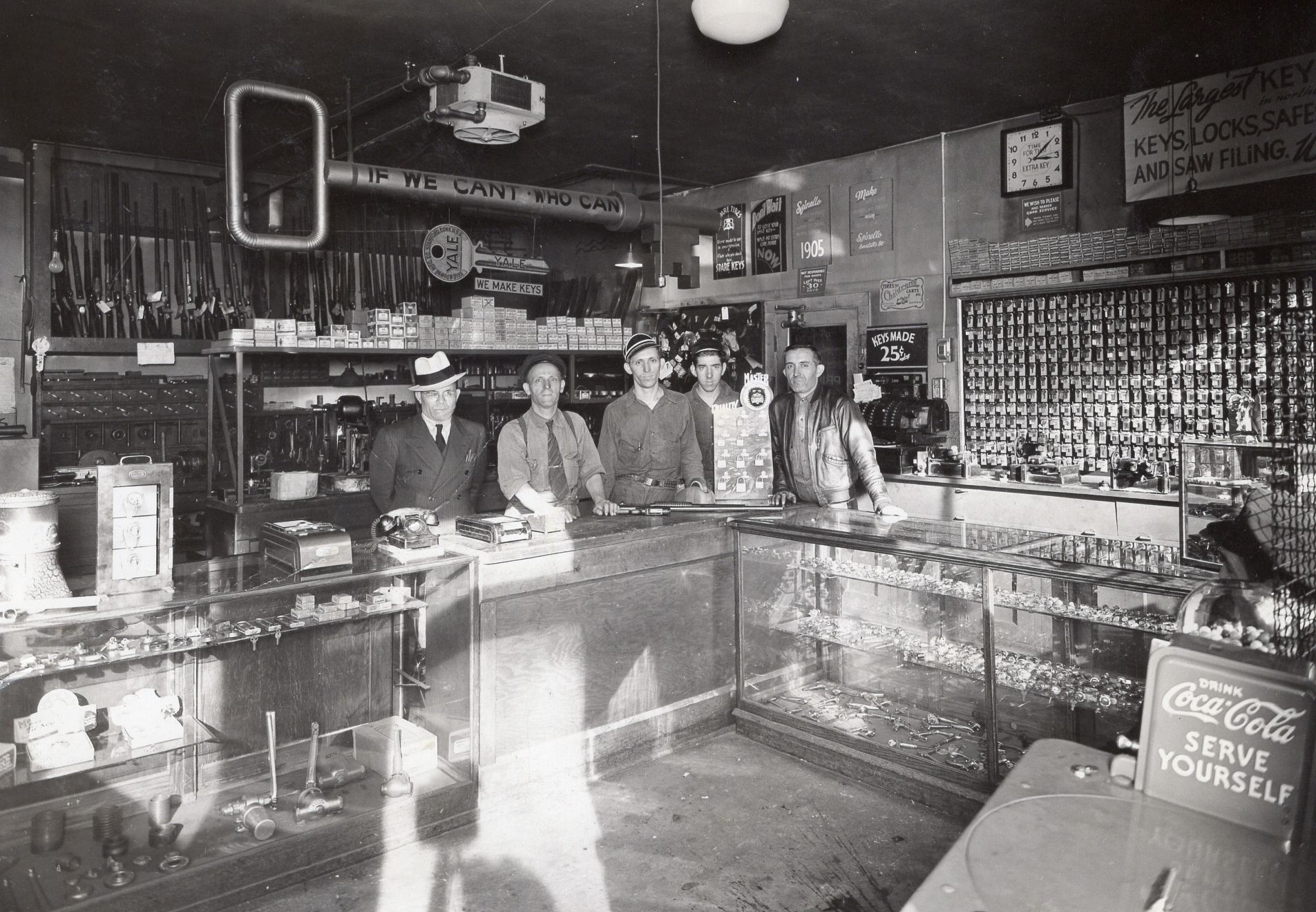 Interior of a shop with several men behind a counter, selling various items, likely hardware.  Coca-cola sign.