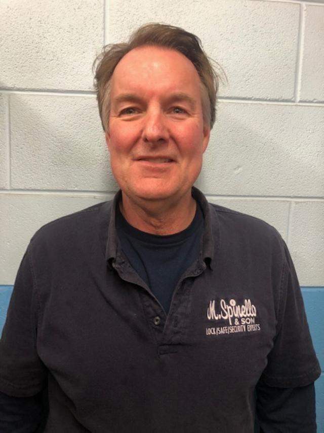 Man in dark shirt with logo, smiling, stands in front of a blue and white wall.