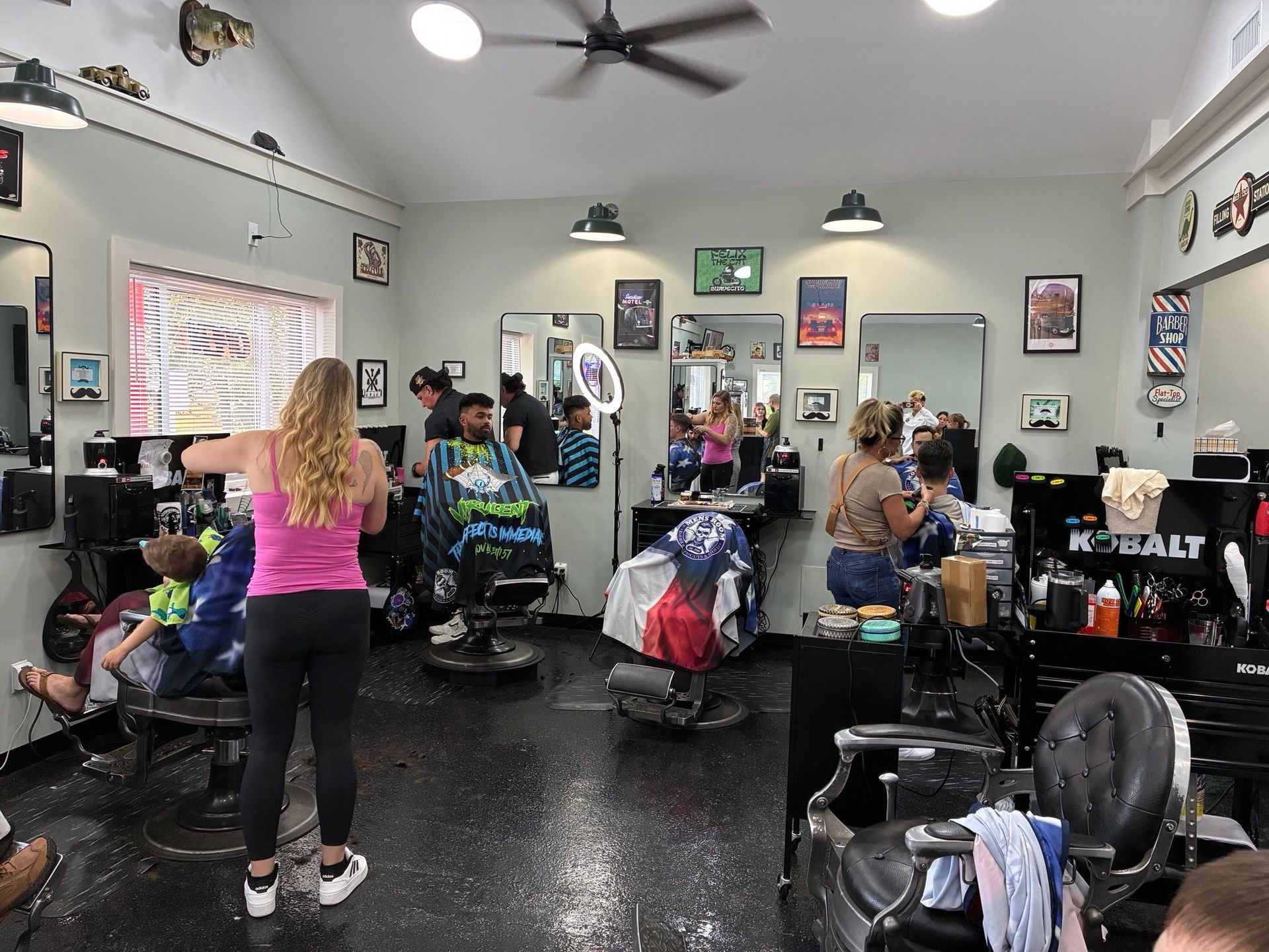 Barbershop with several people getting haircuts, mirrors, chairs, and black-and-white checkered floor.