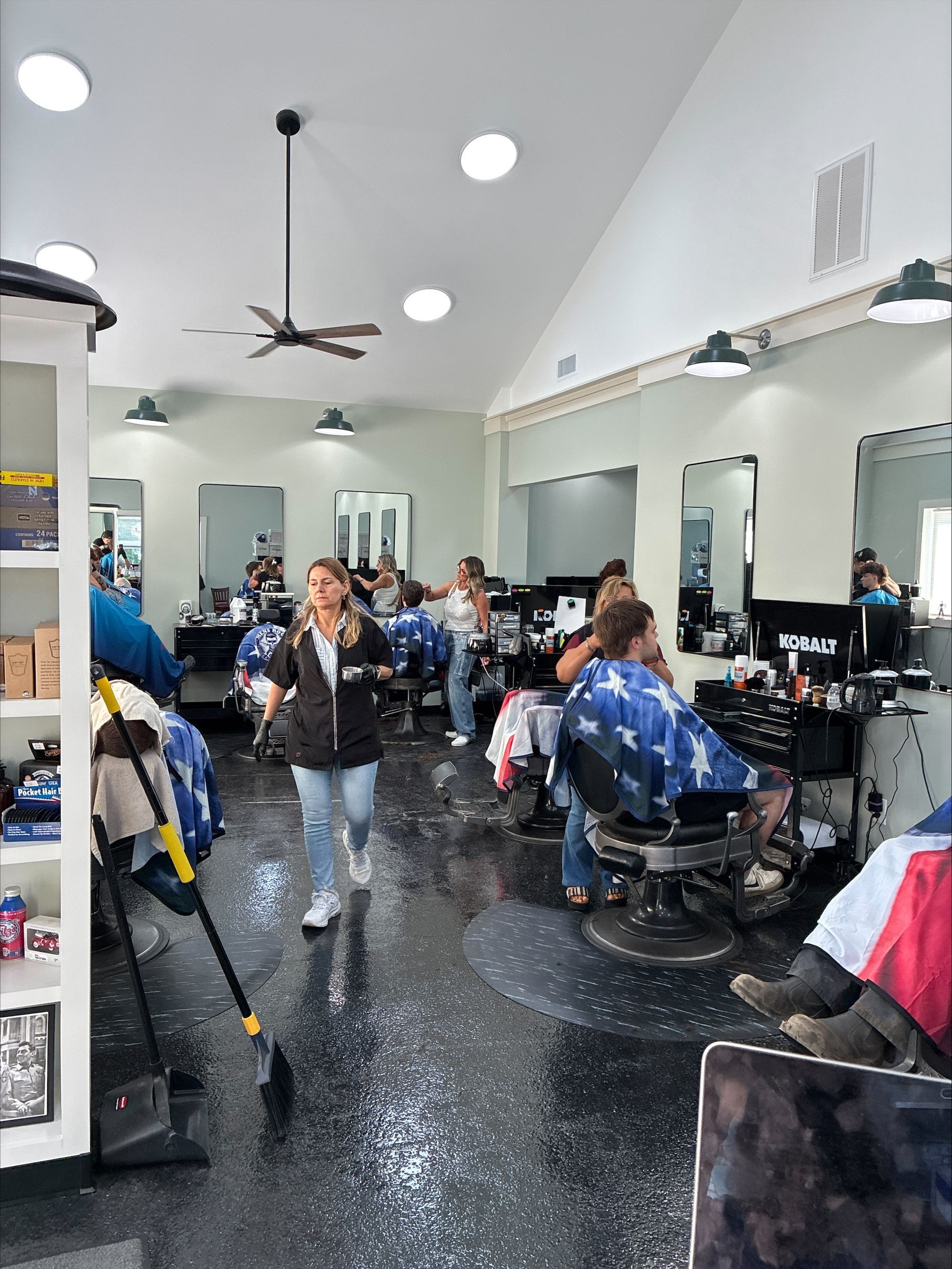 Busy barbershop interior with customers seated for haircuts and stylists working around black chairs.