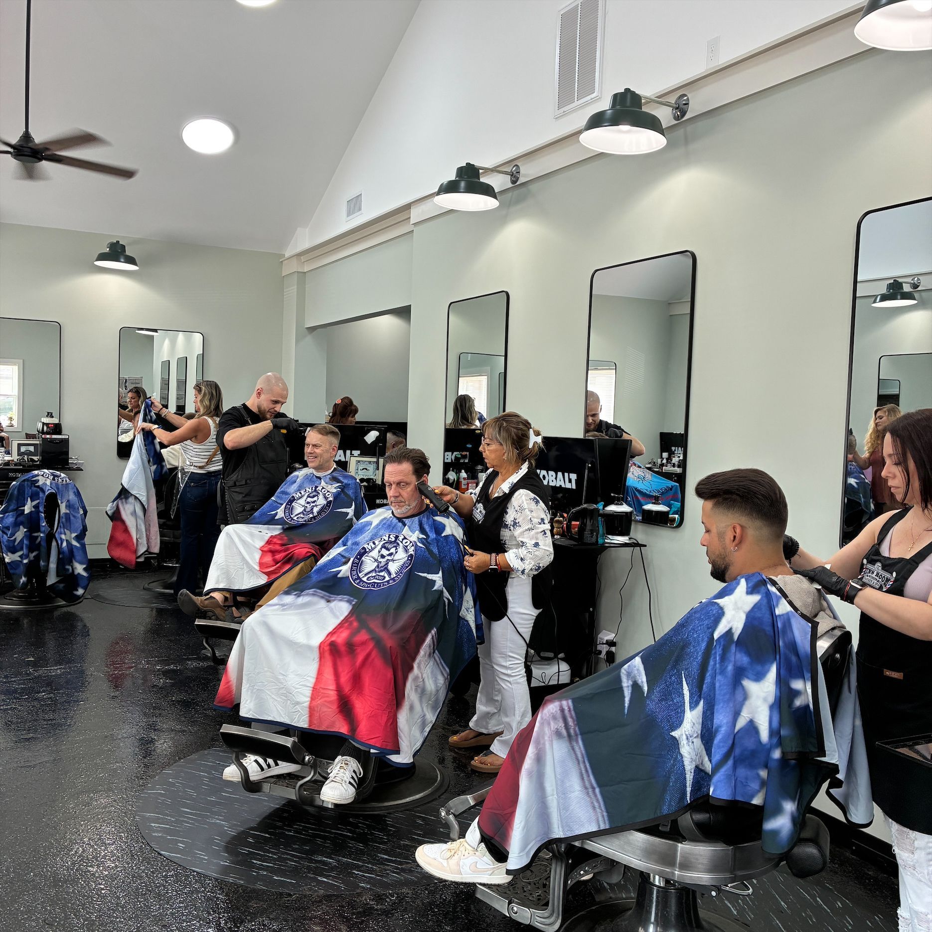 Barbershop with clients in chairs, covered in star-spangled capes, getting haircuts in a bright salon