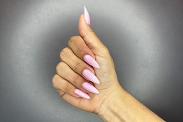 Learn More About Nail Extensions for You Hand with light pink stiletto-shaped nails, raised in a gesture against a gray backdrop.
