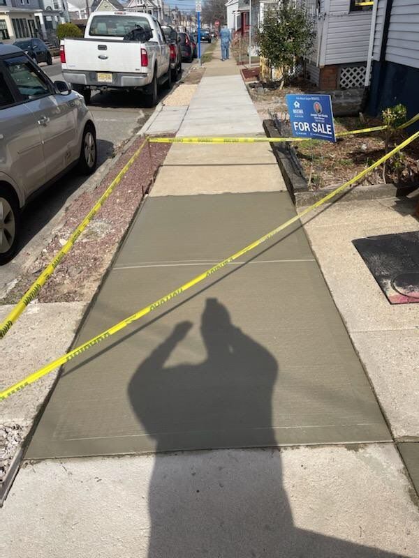 A shadow of a person is cast on a sidewalk next to a for sale sign