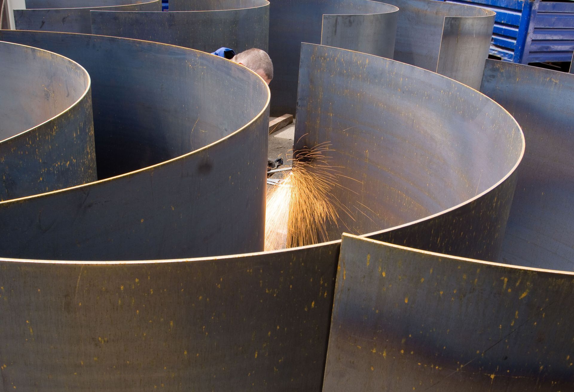 Metal worker cutting curved metal sheets, creating a maze-like structure. Sparks fly from the cutting tool.