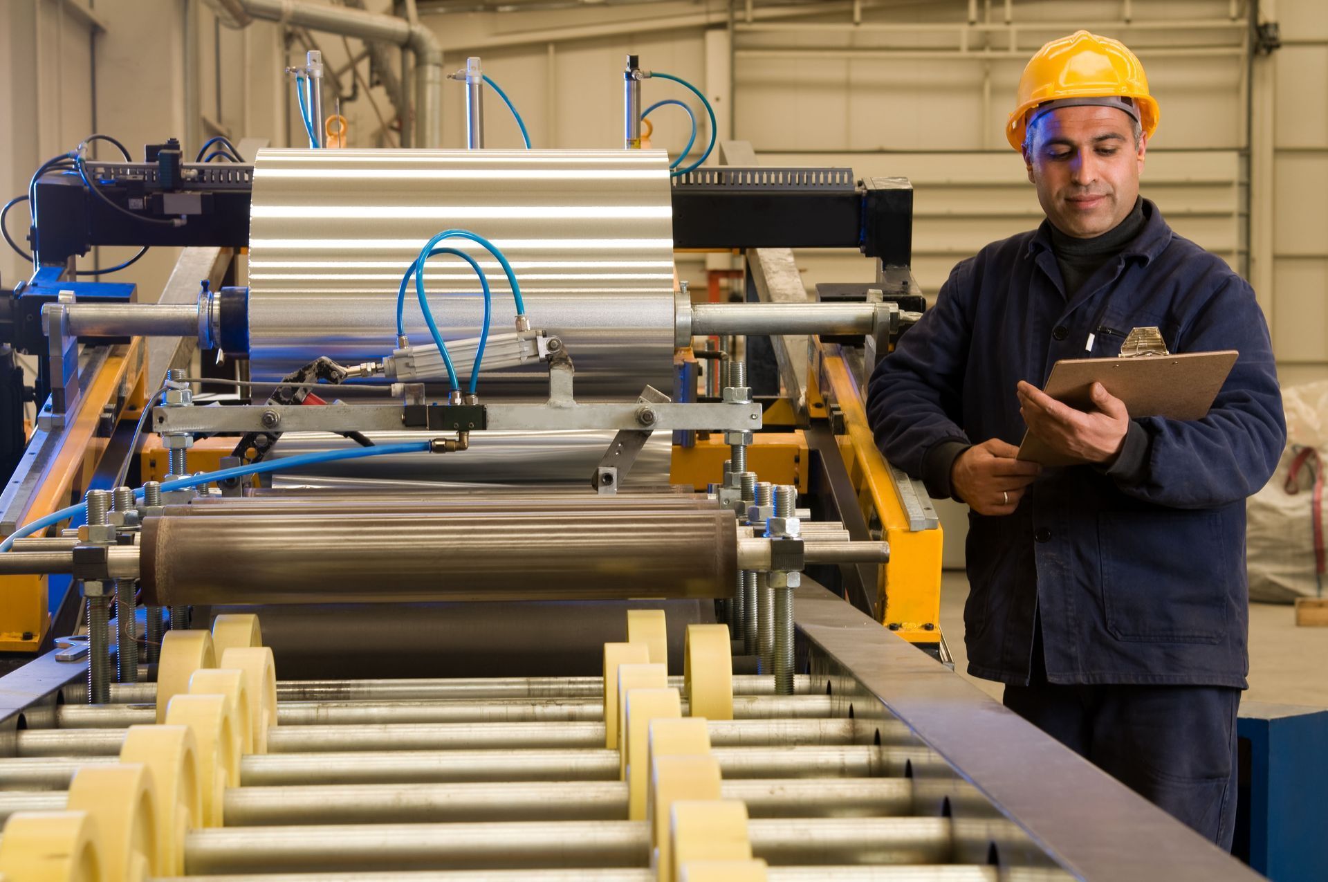 Factory worker in hard hat inspecting machinery with clipboard.