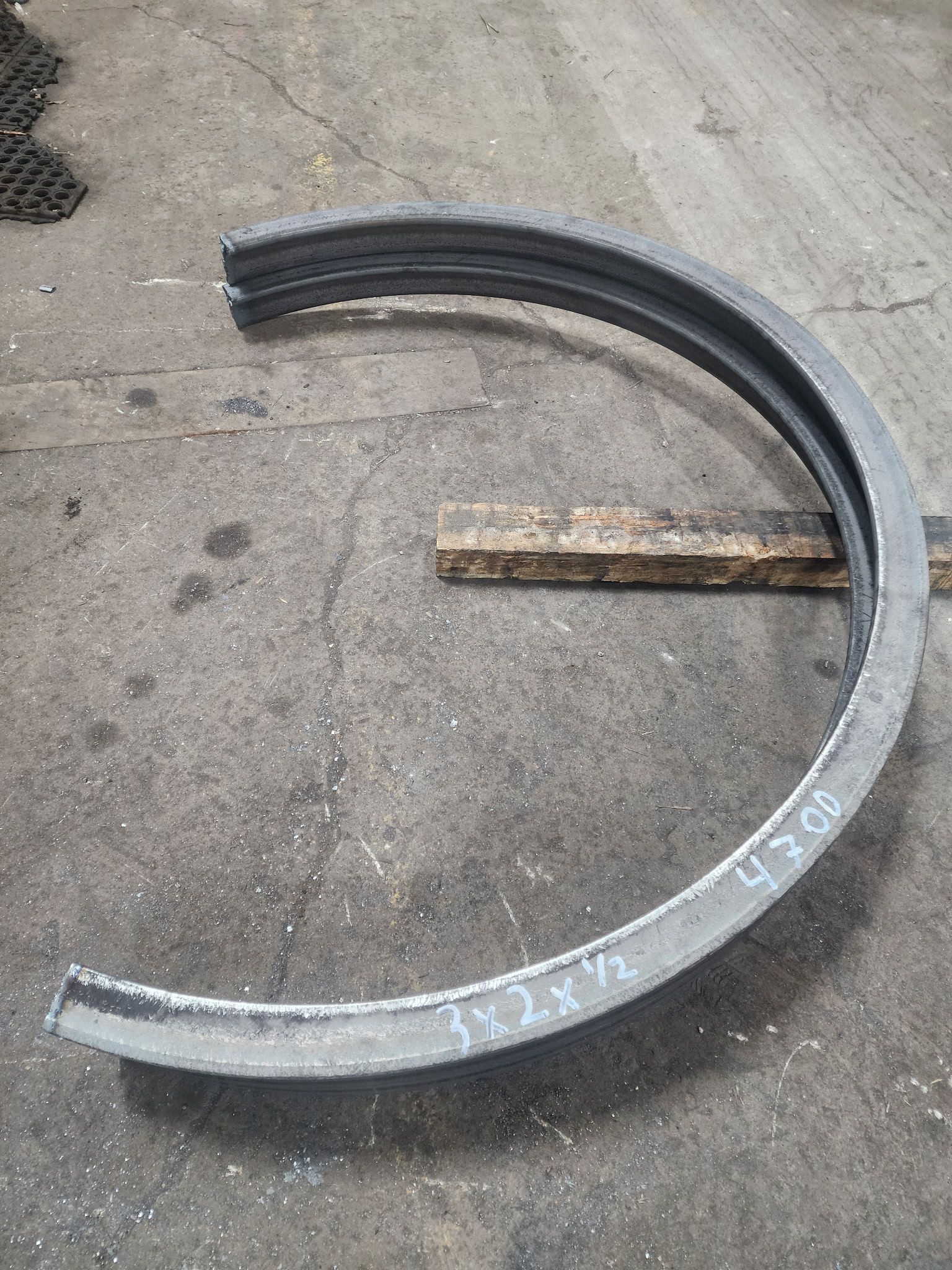 A curved, dark metal industrial angle iron component resting on a concrete floor with hand-written measurements.