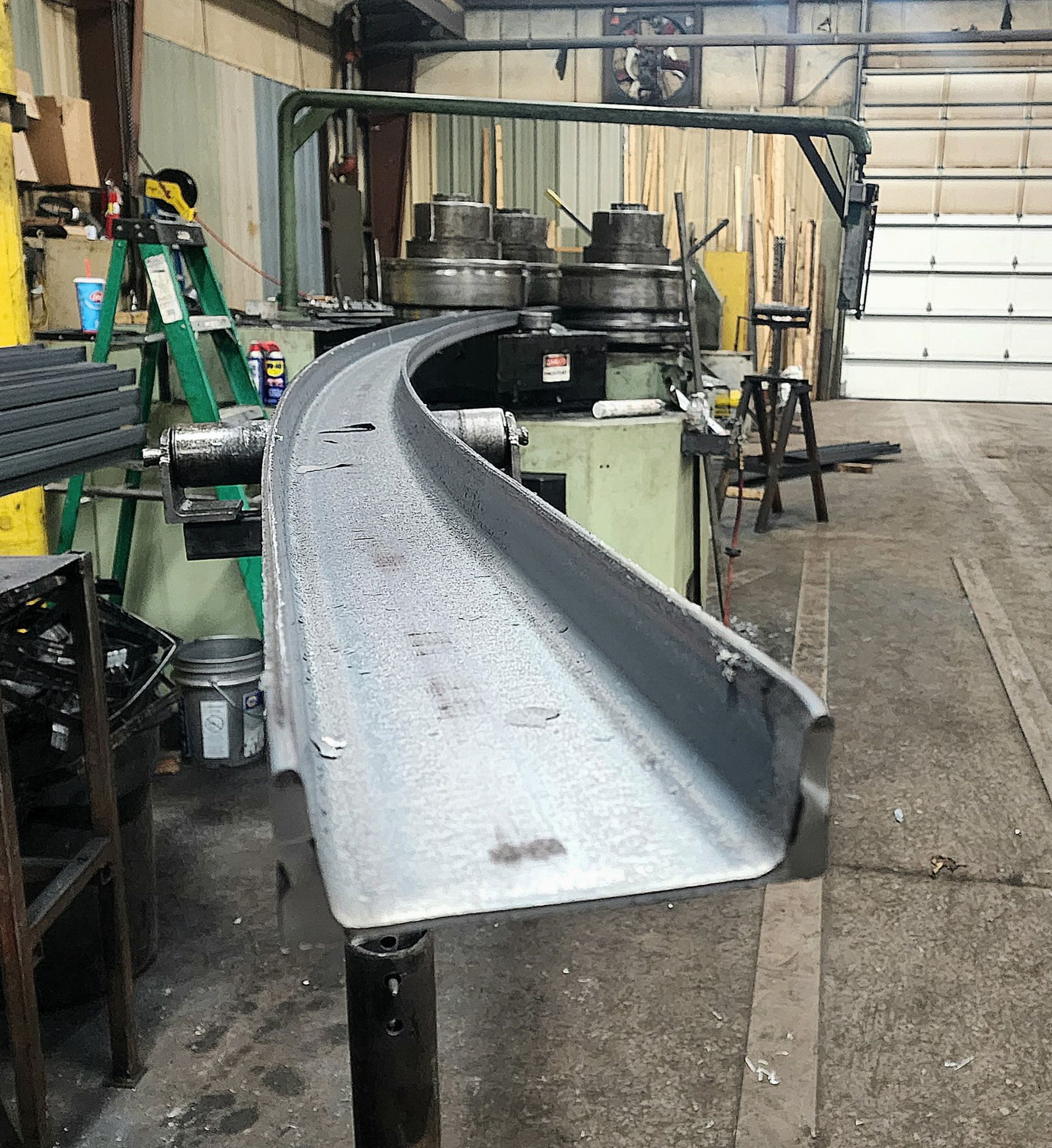 A curved metal beam in a workshop setting, possibly for construction, near machinery.