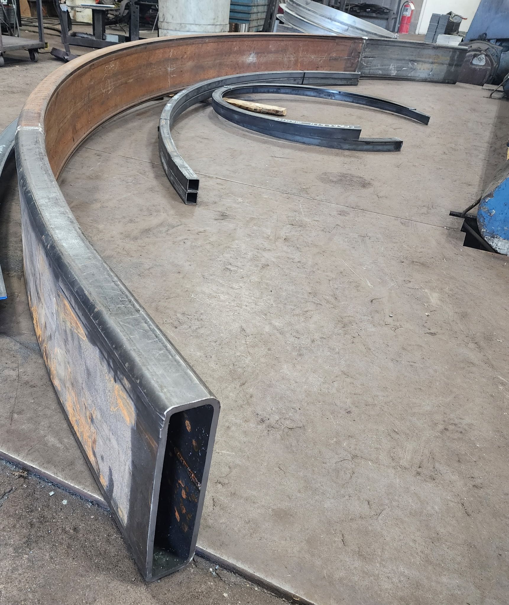 Curved metal beams on a workshop floor; some are rectangular, others in arcs, with a rusty hue.