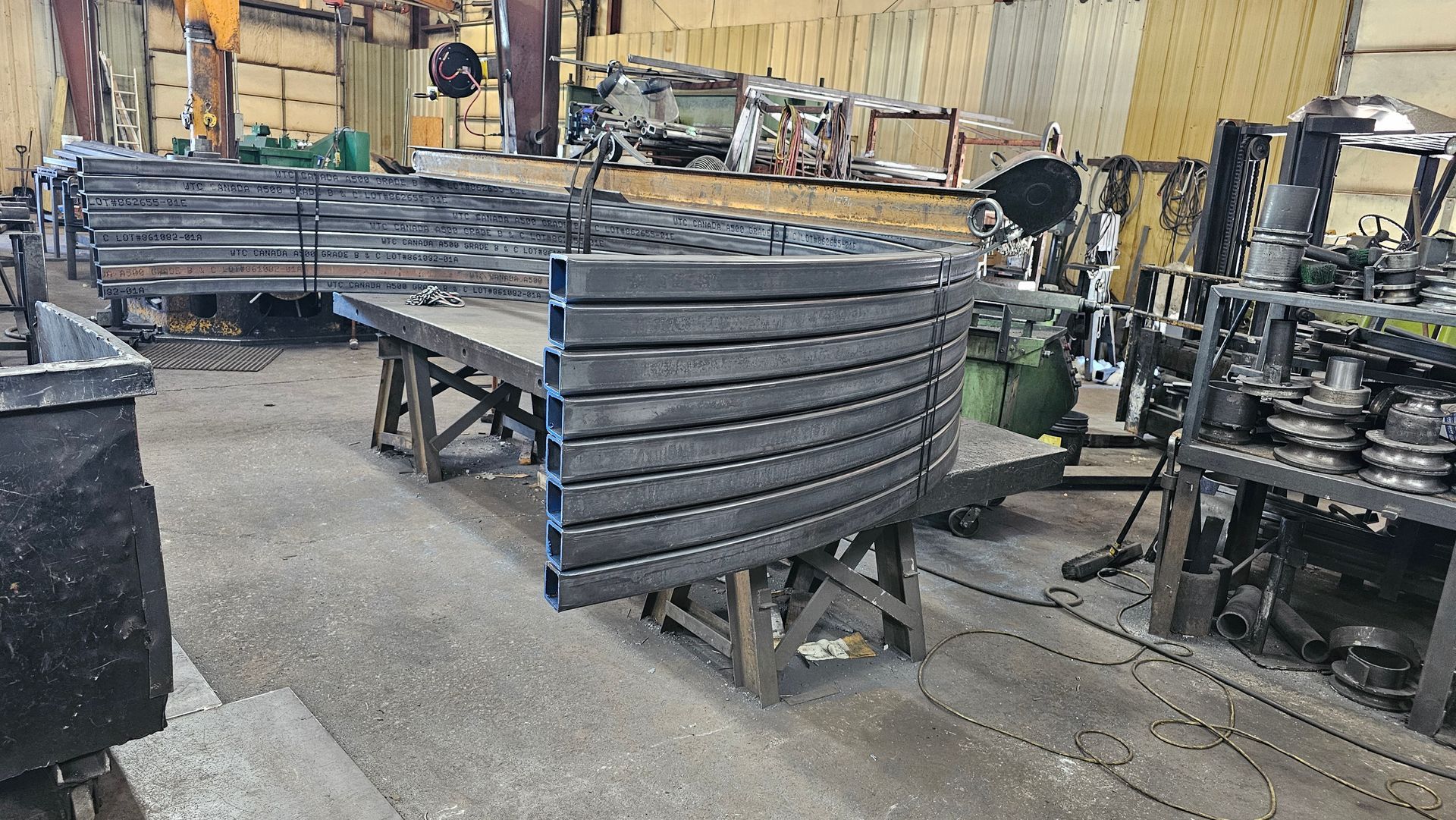 Metalwork in a workshop: Curved steel bars stacked on a table. Various equipment and materials in the background.
