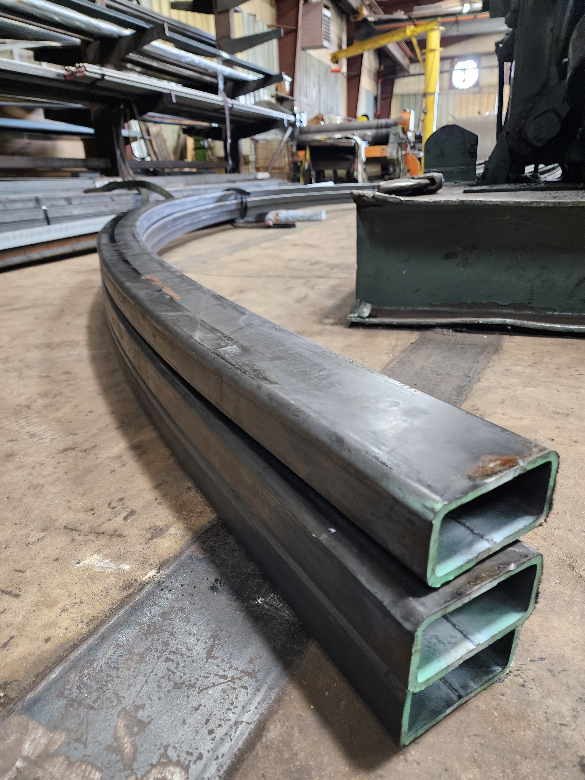 Two curved steel rectangular tubes in a workshop, stacked and lying on a metal surface.
