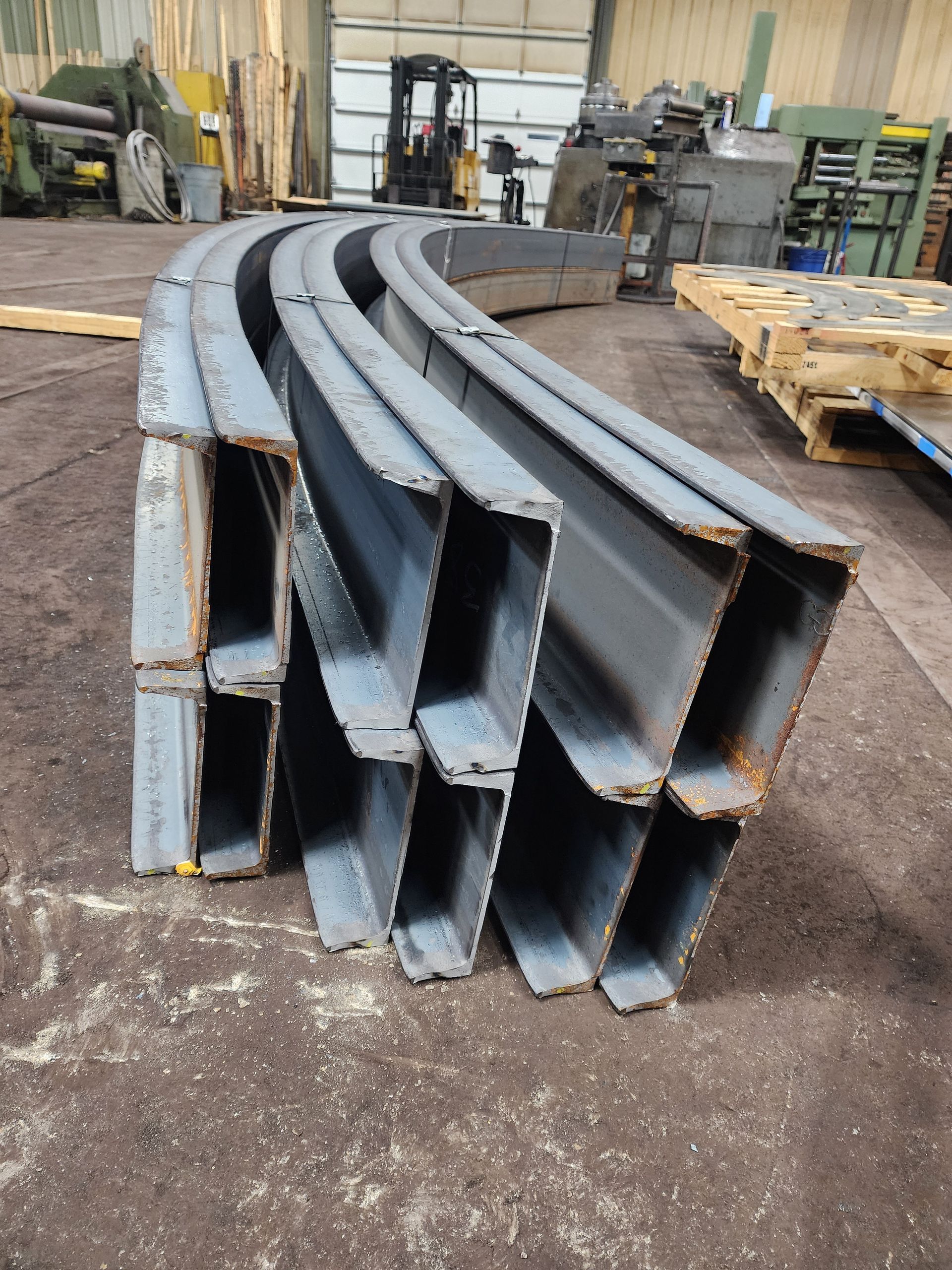 Curved steel beams stacked on shop floor. Industrial setting with equipment and forklift in background.