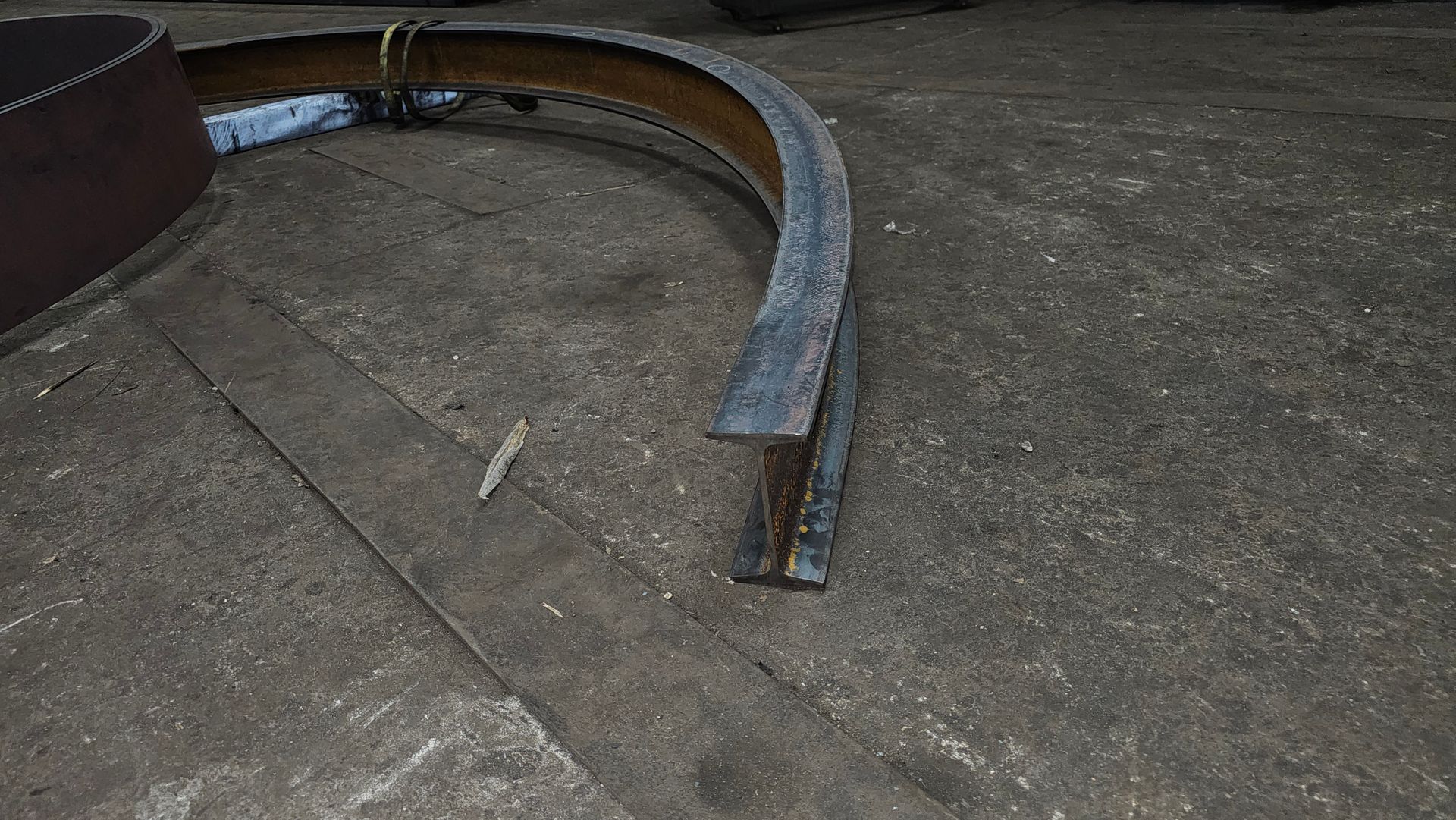 Curved steel beam section on a concrete surface, near a larger cylindrical metal structure.