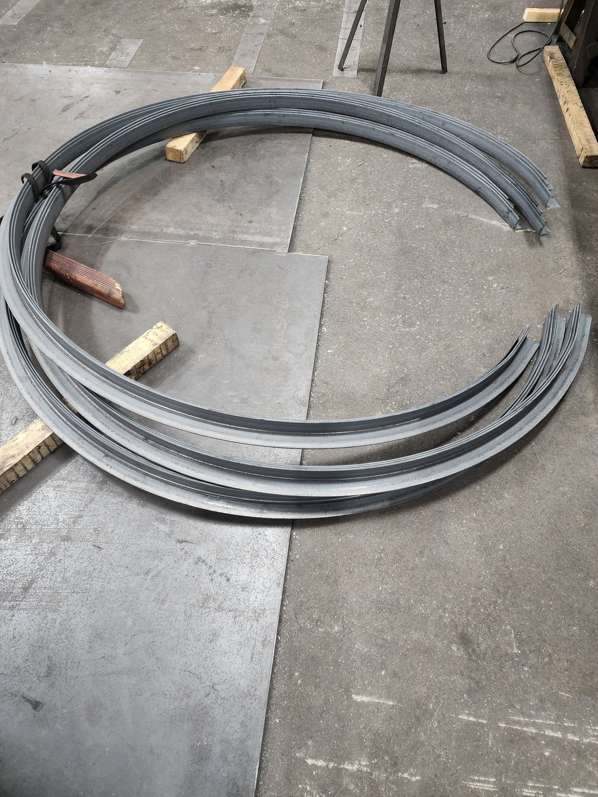 Metal curved beams stacked on a concrete floor with wooden supports.