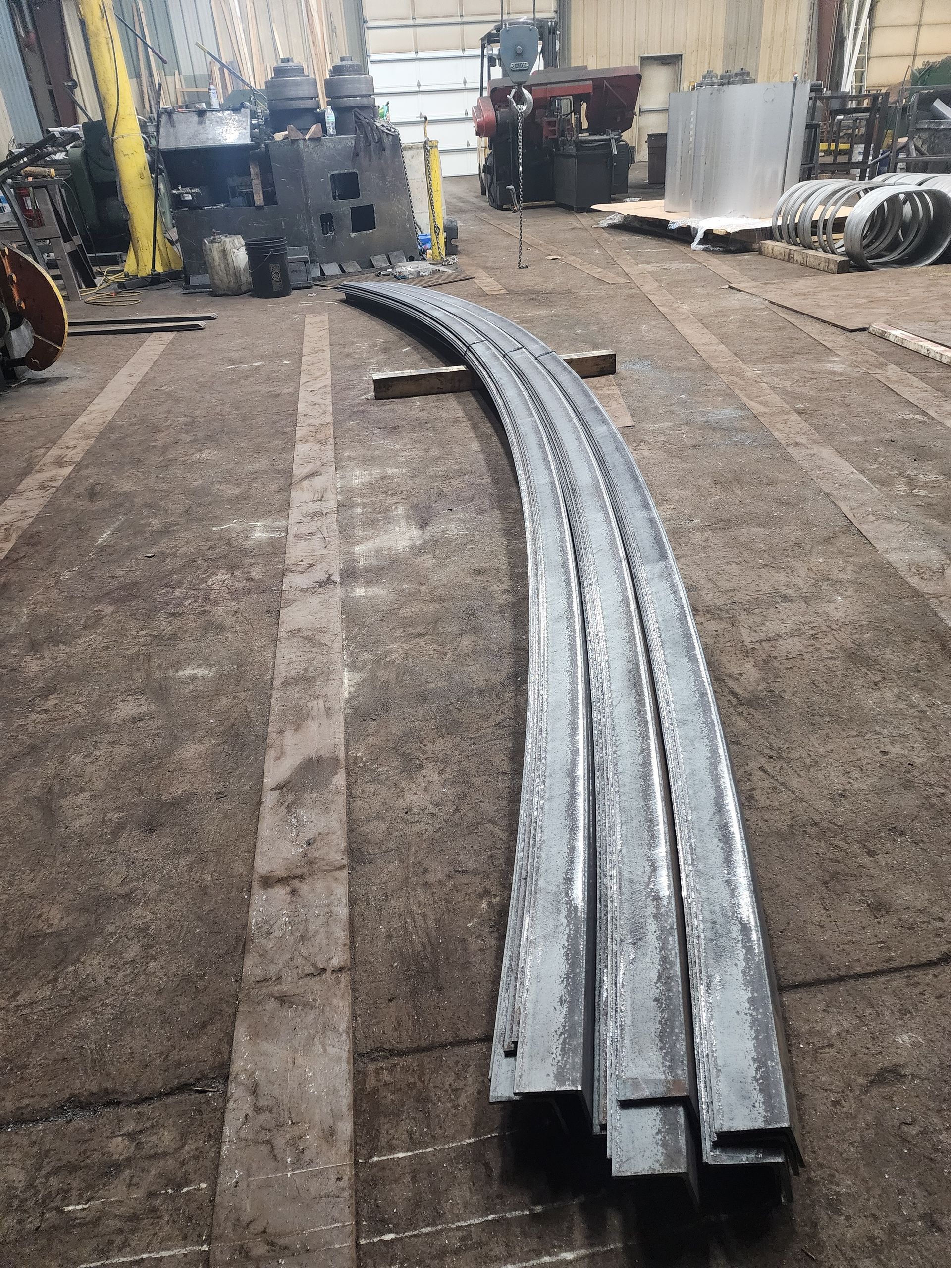 Curved metal beams on a factory floor. Background shows machinery and other equipment.