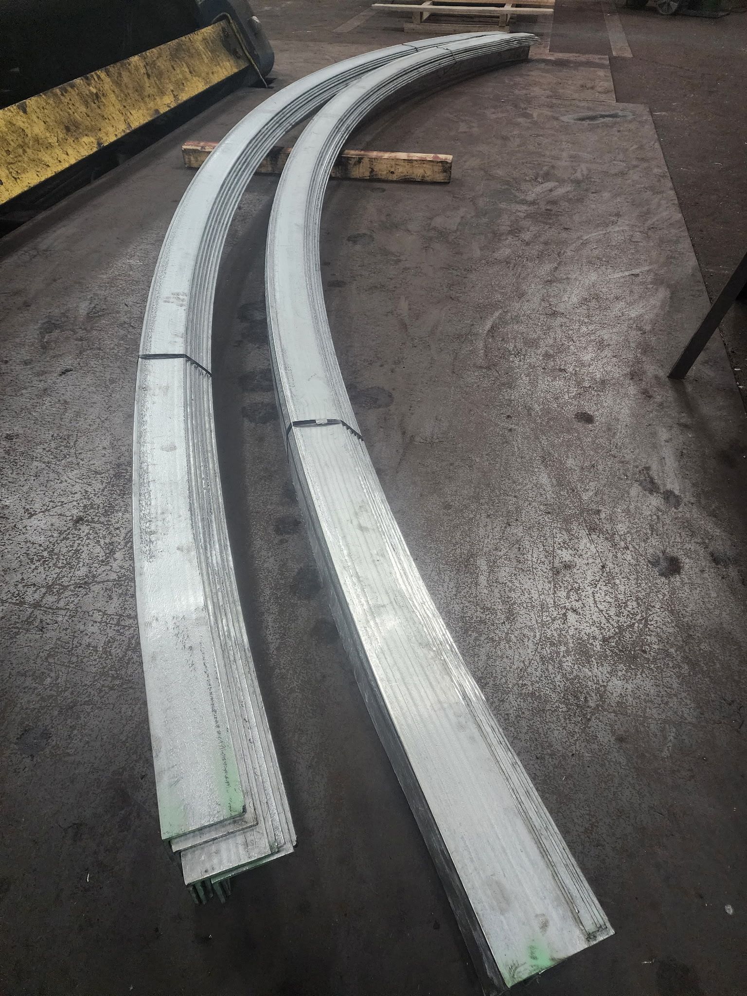 Two stacks of curved, galvanized steel beams on a concrete floor.