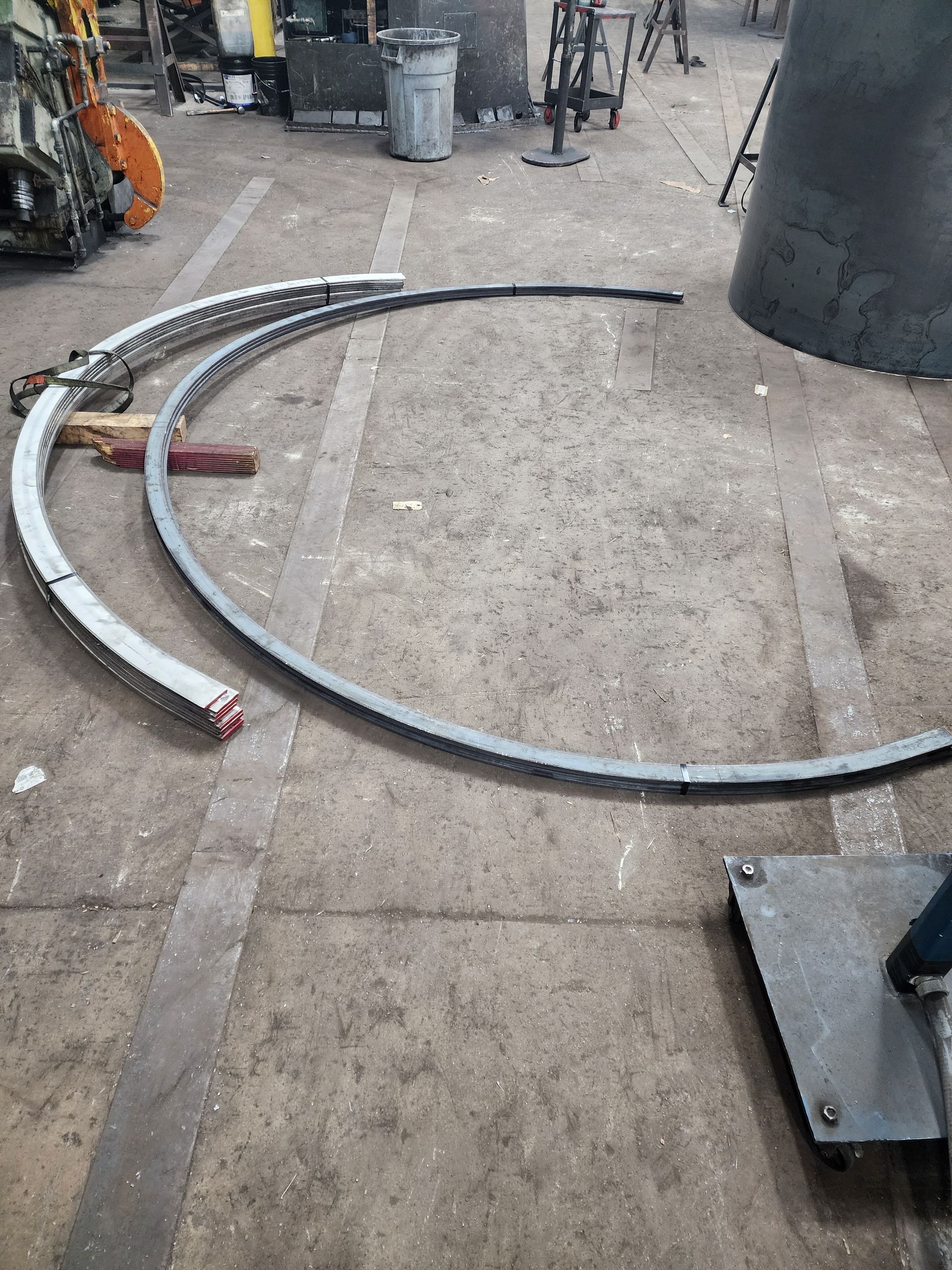 Curved metal pieces arranged on a shop floor near a cone-shaped structure and other equipment.