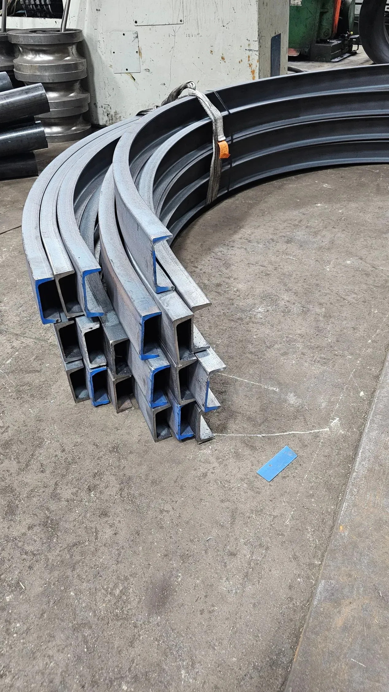 Curved metal beams, stacked. Blue tape on ends. Industrial setting.