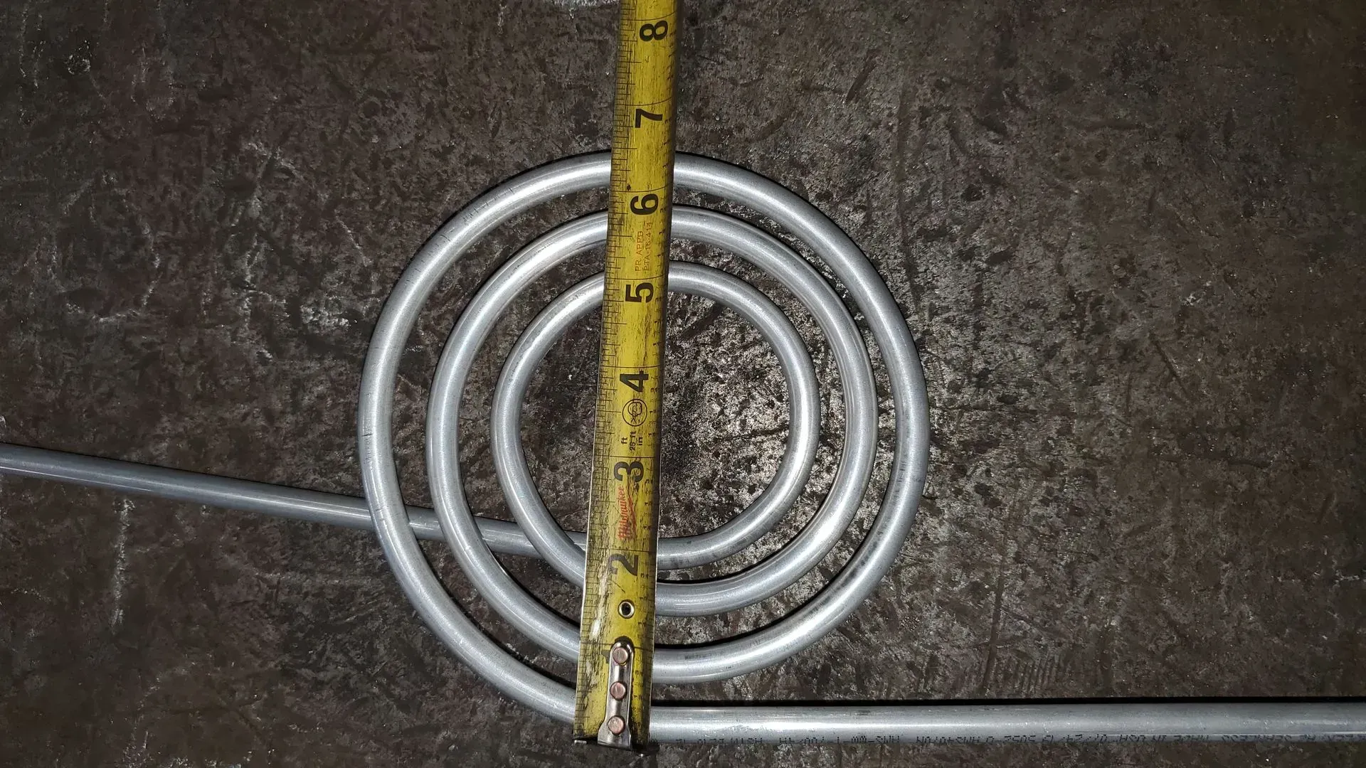 Coiled metal tubing with a yellow tape measure, on a metal surface.