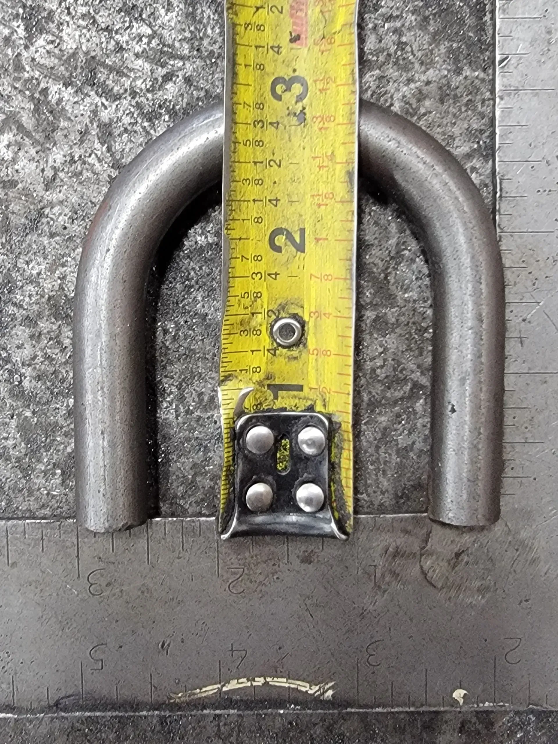 A metal U-shaped object is measured with a yellow tape measure on a metal surface.