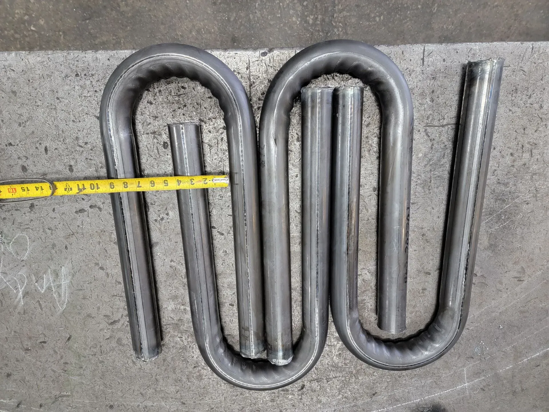 Bent metal tubing in an S-shape, measured by a yellow tape measure, on a metal surface.
