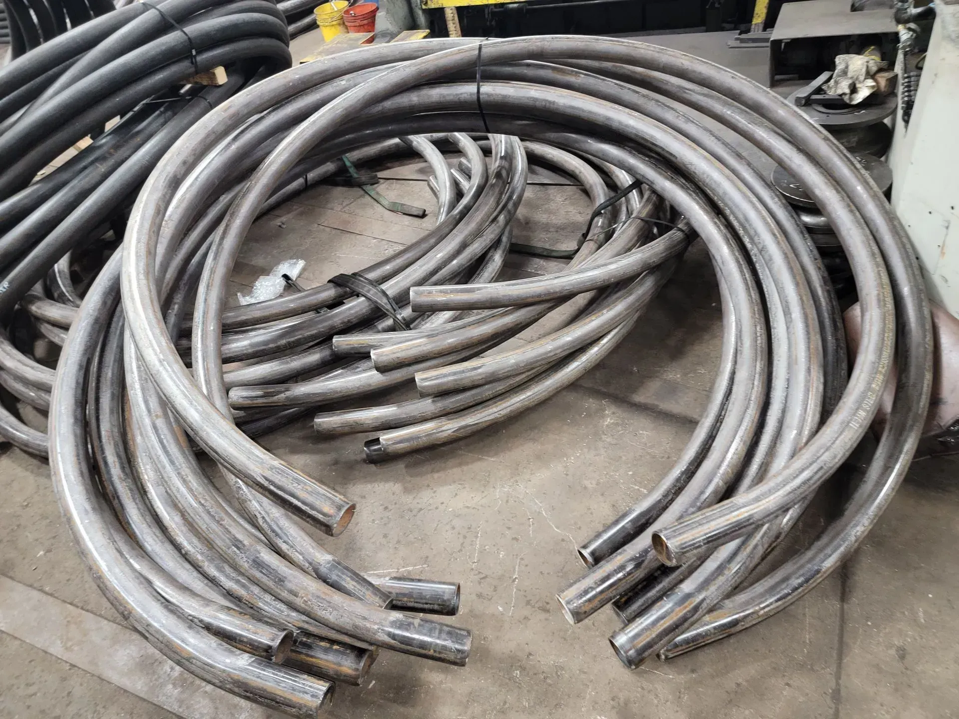 Curved metal tubes, silver, piled on a surface, possibly for construction or manufacturing.