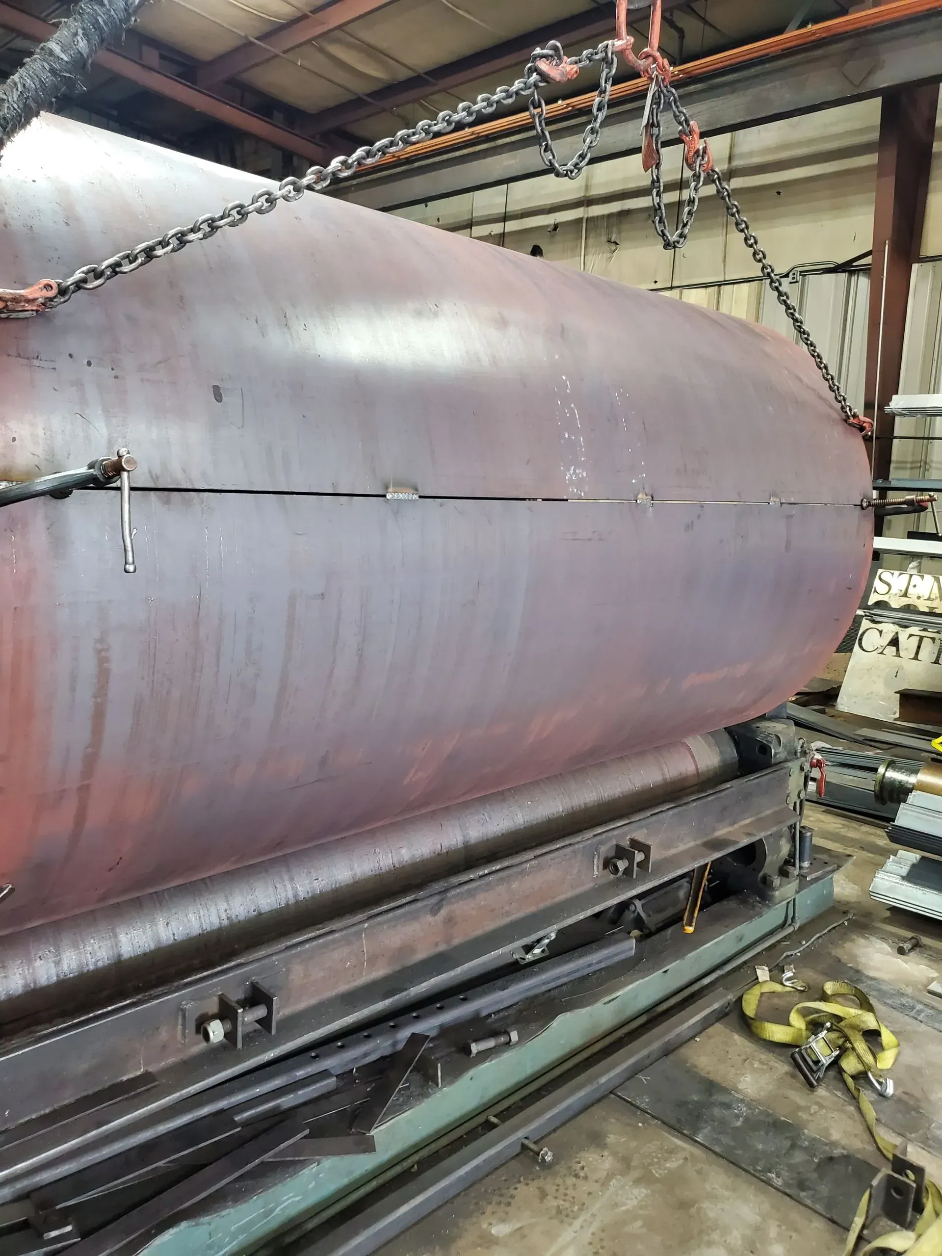 Large, reddish-brown metal vessel suspended by chains above a machine in a workshop.